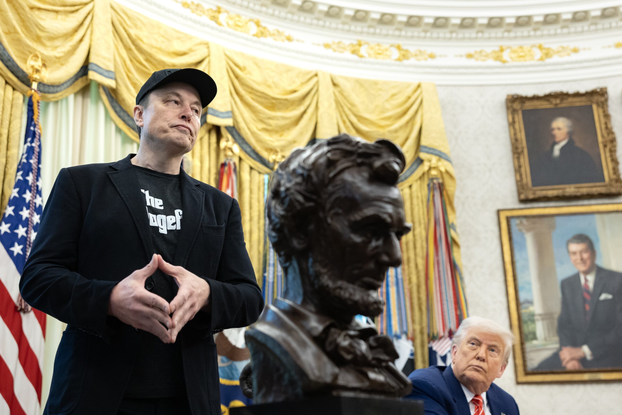 Elon Musk stands in the oval office next to Trump, who stares up at him. Between them is a bust of Abraham Lincoln.