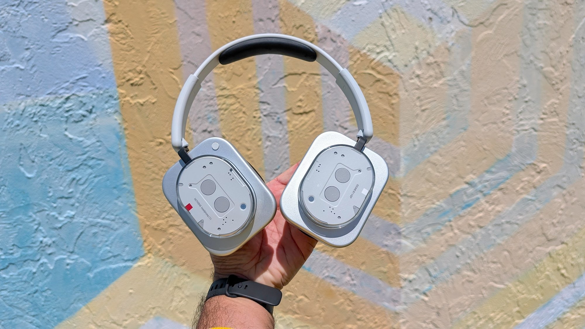 man's hand holding up a pair of Nothing headphones against mural