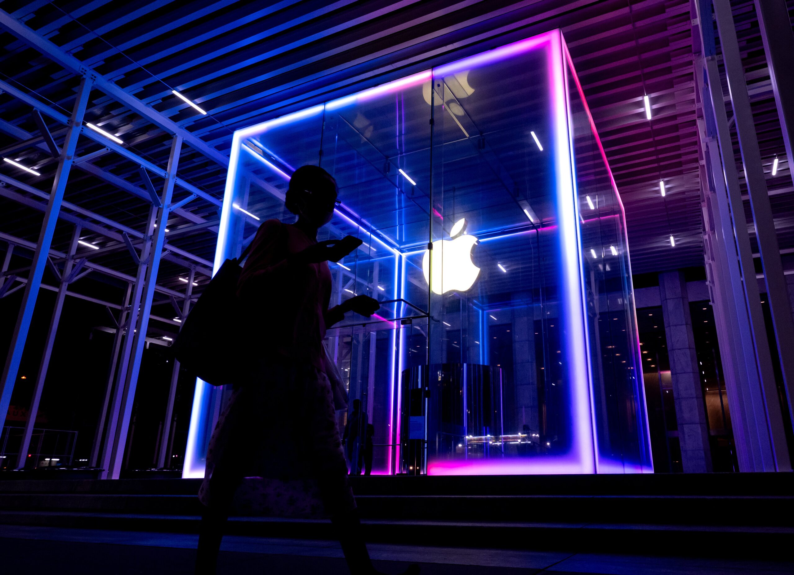 A glowing multi-colored glass cube in a dark room. A glowing Apple logo sits in the middle as a person in shadow walks by.