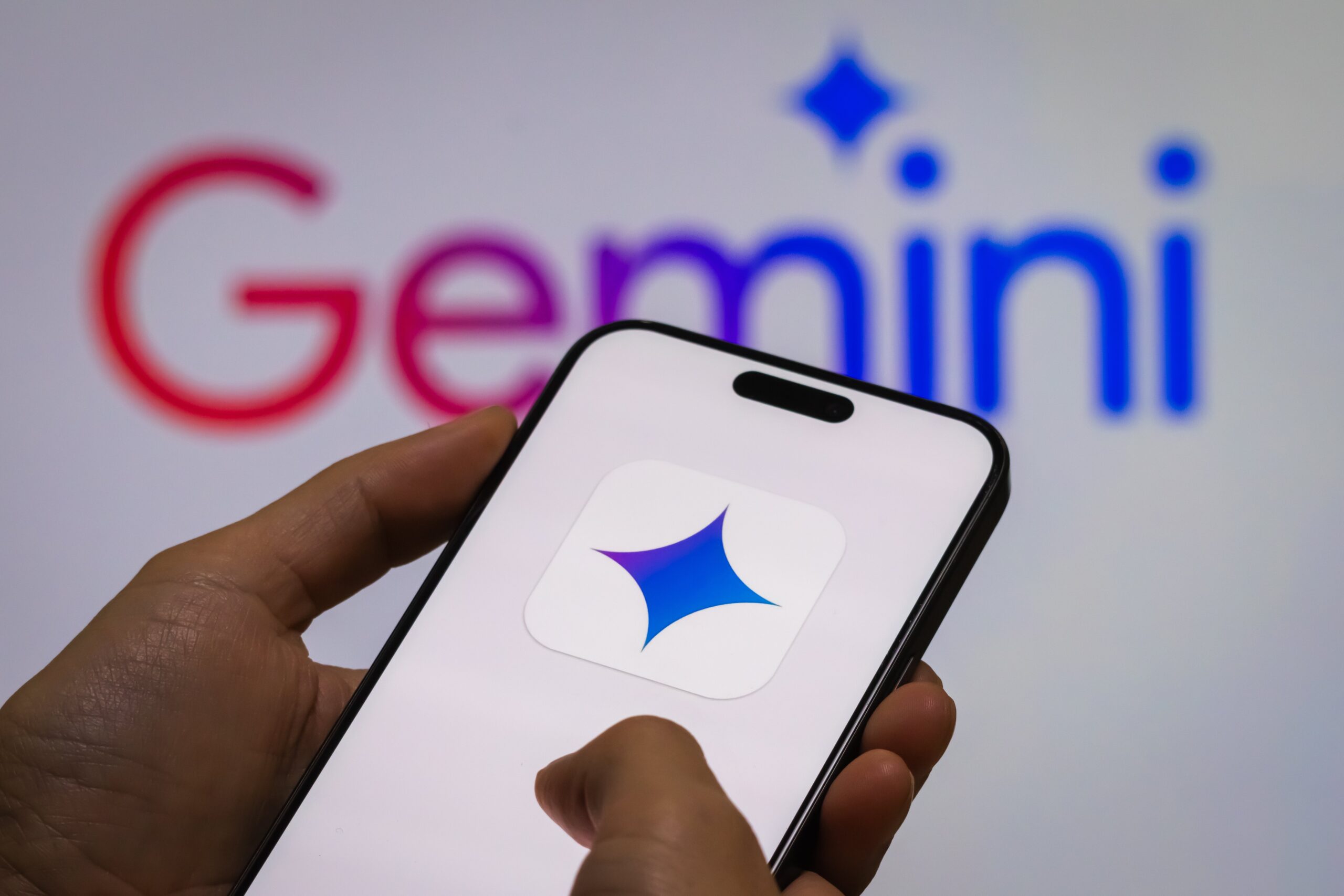 a person holds a smartphone displaying the logo of “Gemini,” Google’s next-generation AI model, with the “Gemini” logo visible in the background