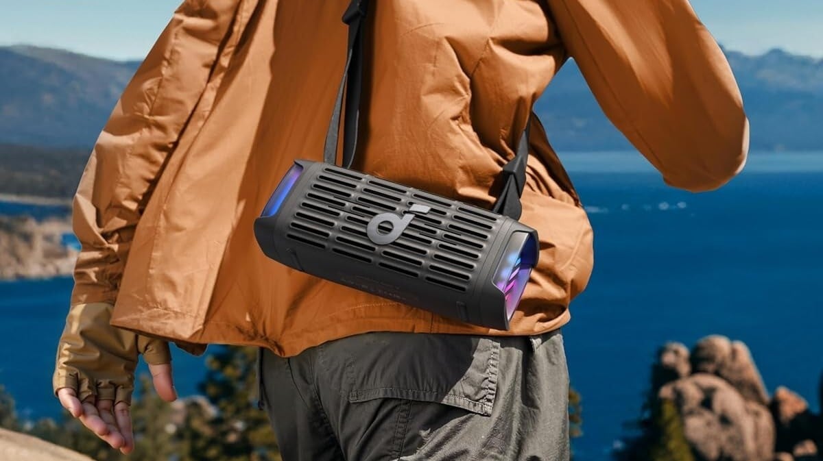 a person wearing an orange rain shell holds the soundcore boom 3is speaker slung over their shoulder while walking toward a body of water