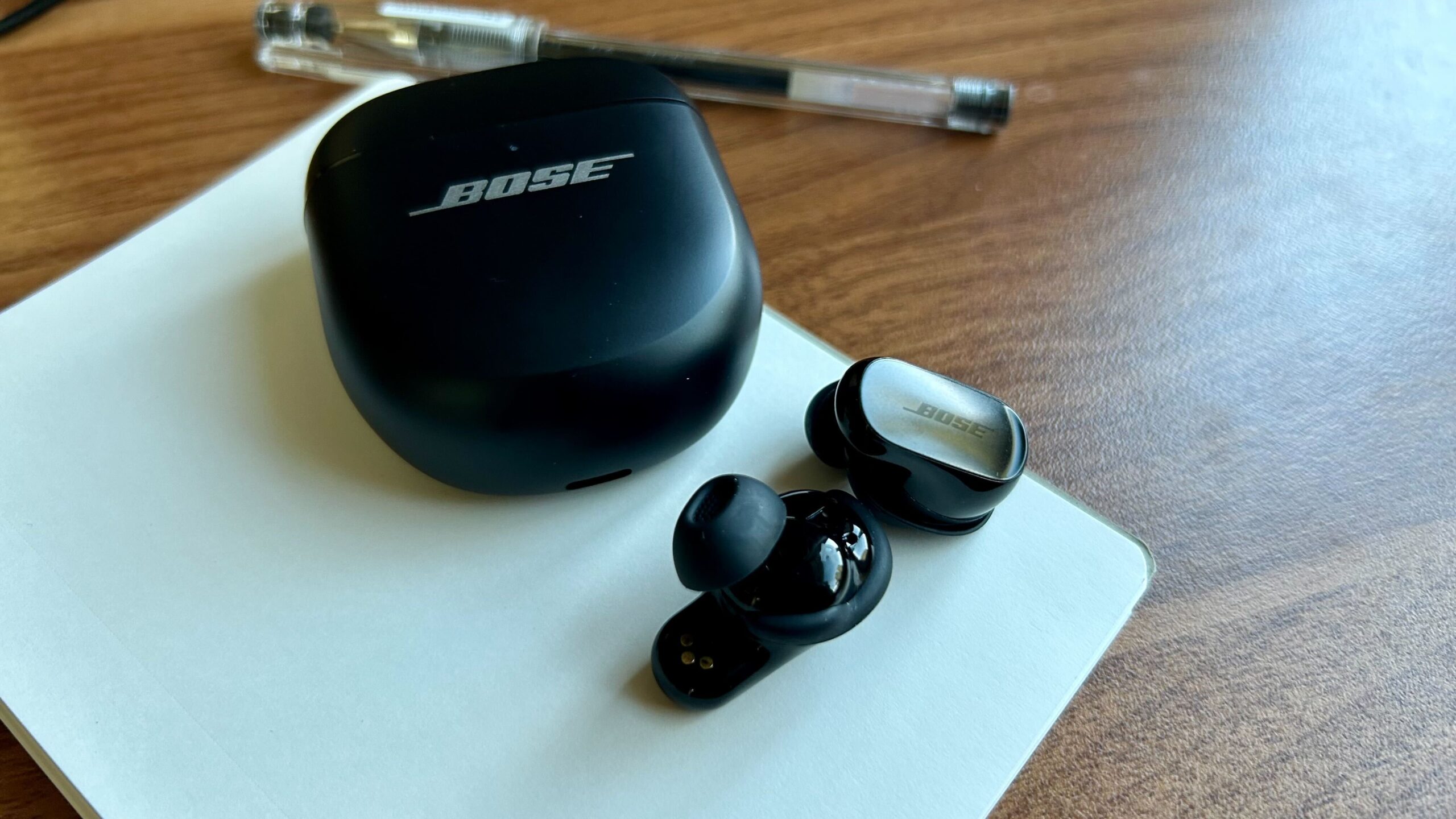 black bose qc ultra earbuds on notebook near pen on desk