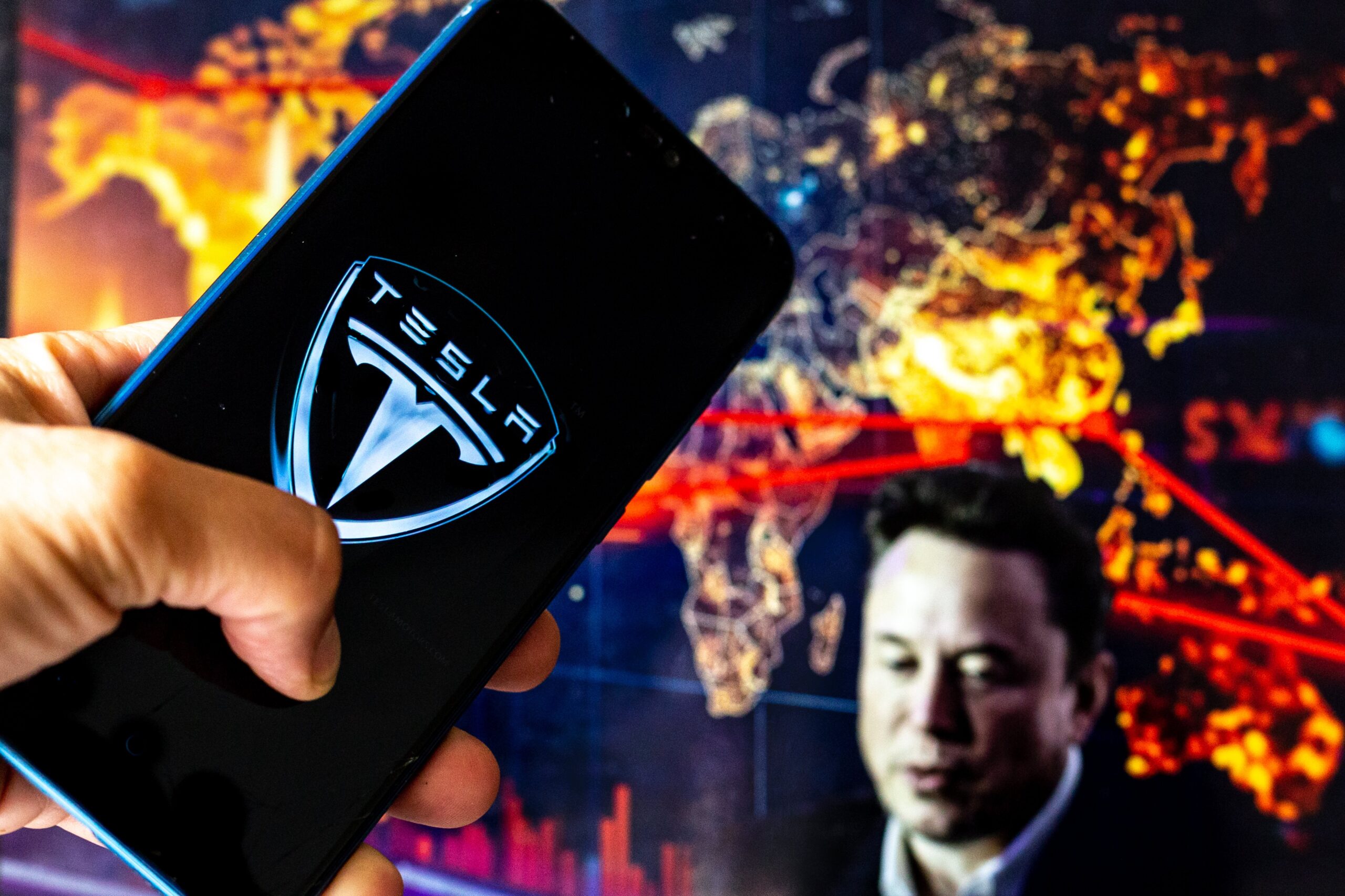 Tesla logo on mobile device with Elon Musk in the background