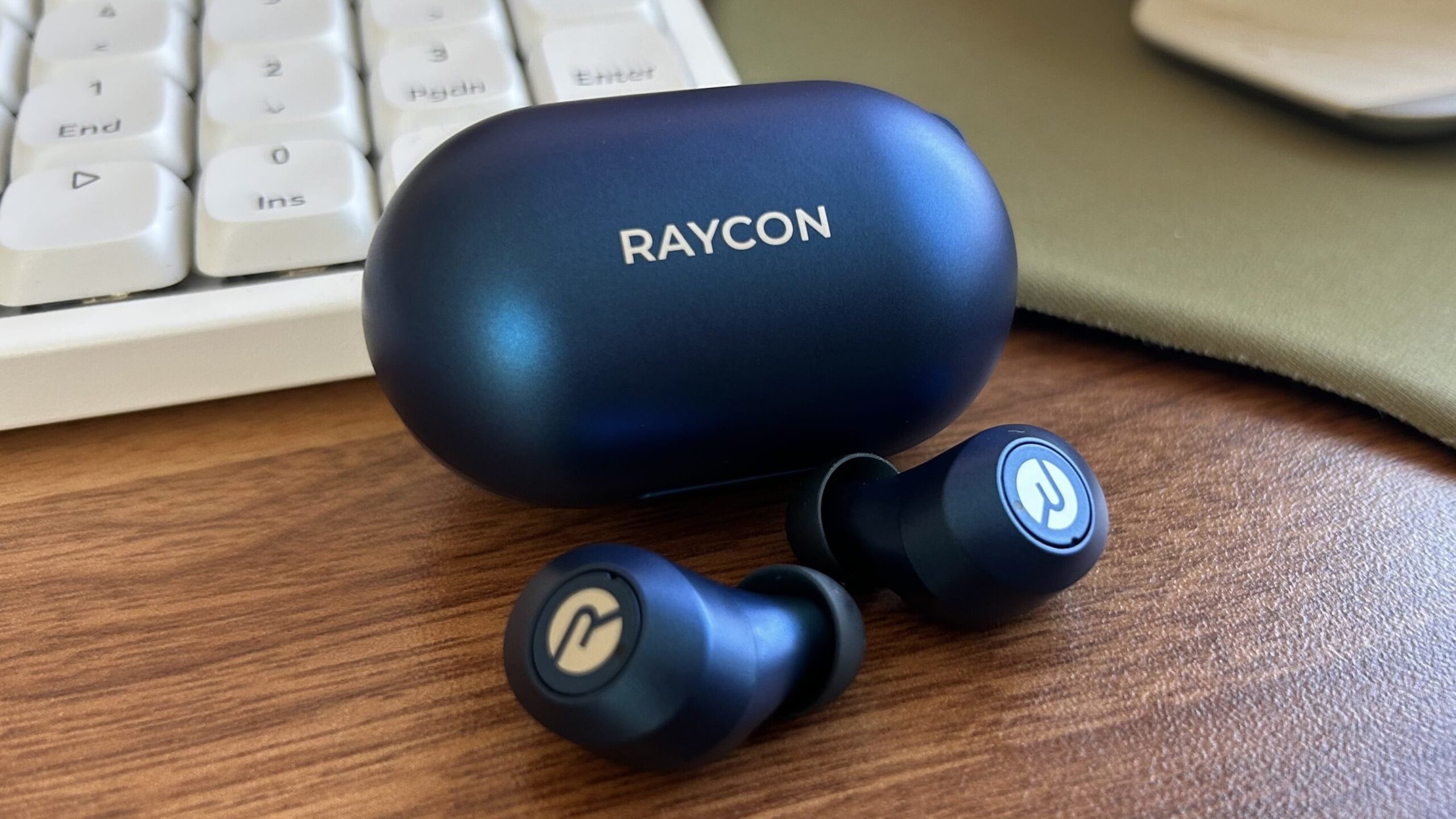 blue raycon earbuds on desktop next to white keyboard and green mouse pad