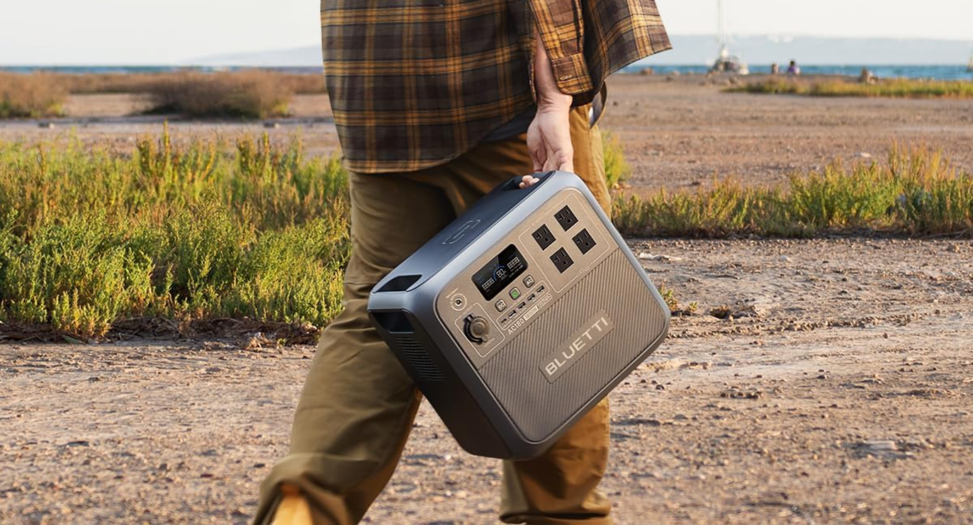 A person in a plaid shirt carries a gray Bluetti portable power station by its handle while walking outdoors in a grassy, coastal area.