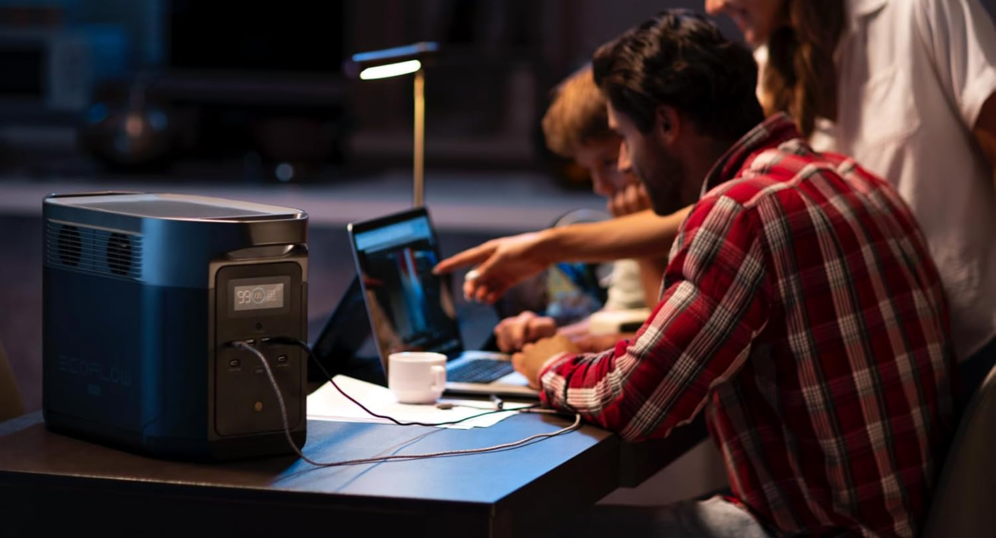 An EcoFlow portable power station sits on a wooden desk, powering a lamp and a laptop while three people work together in a dimly lit room.