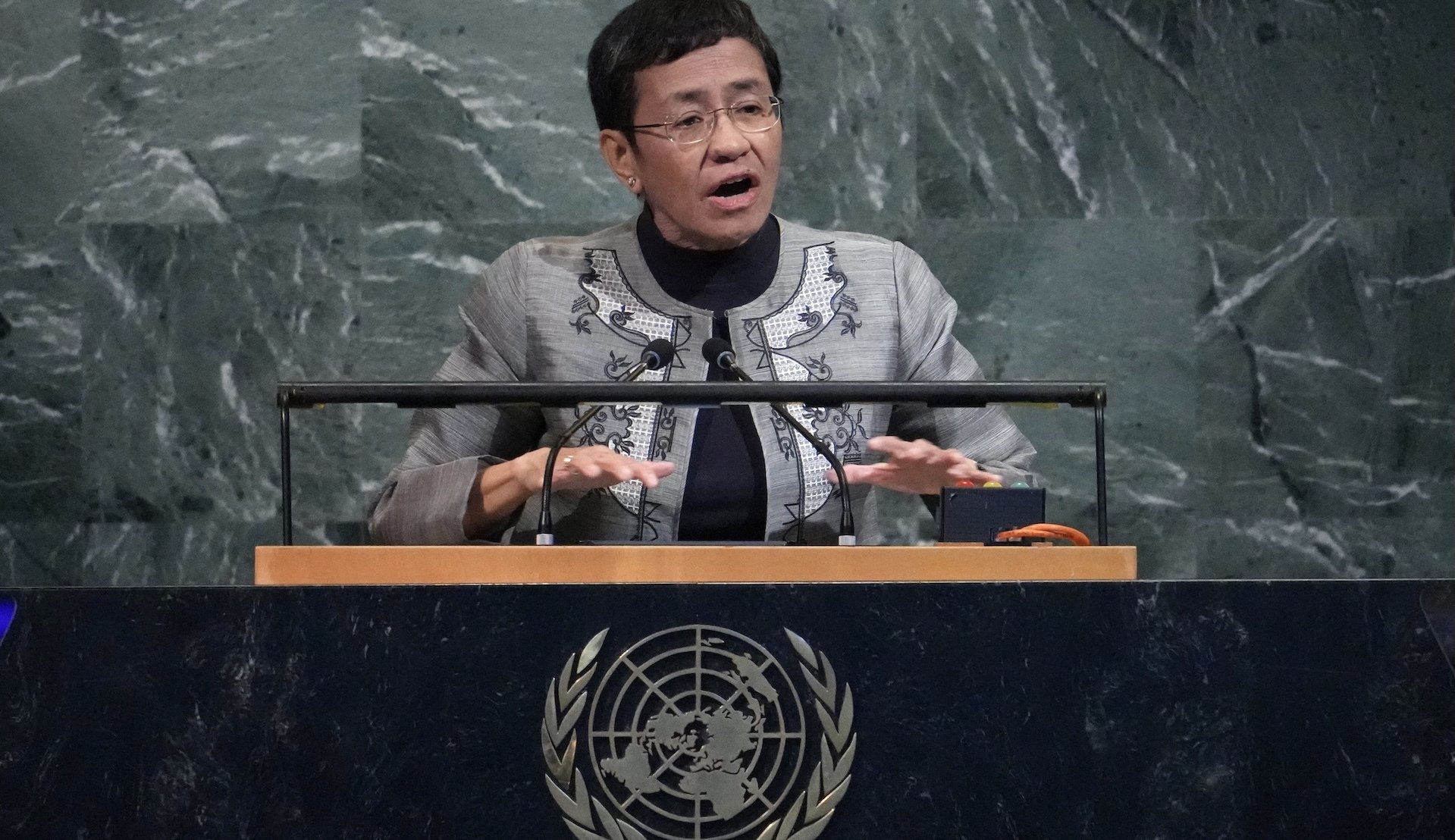 At the UN podium, a woman with short hair and glasses speaks. 