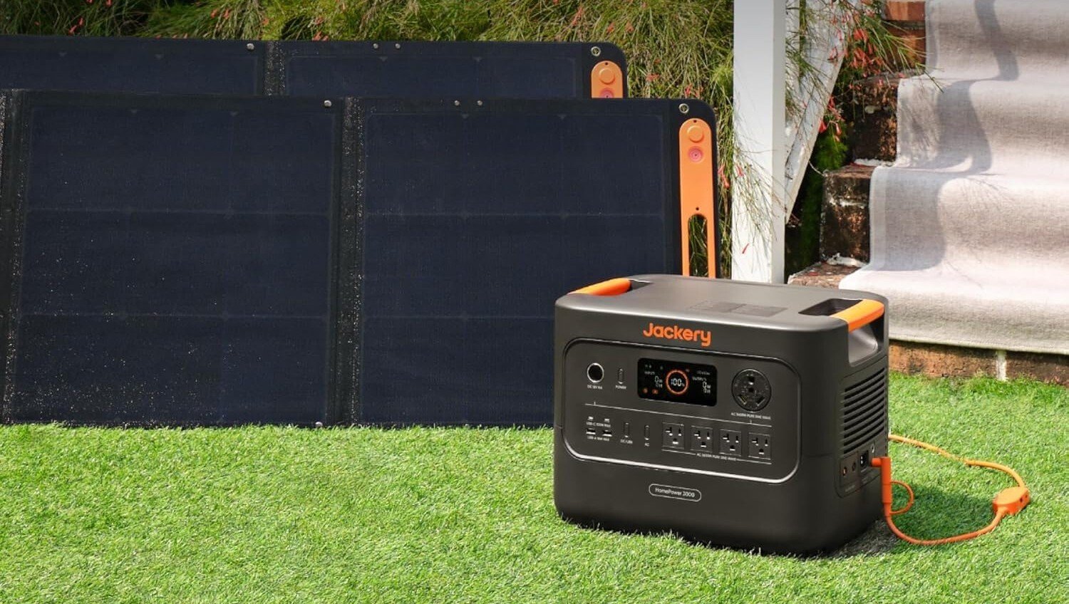 the jackery homepower 3000 sits in the grass on a sunny day while two solar panels sit behind the portable power station