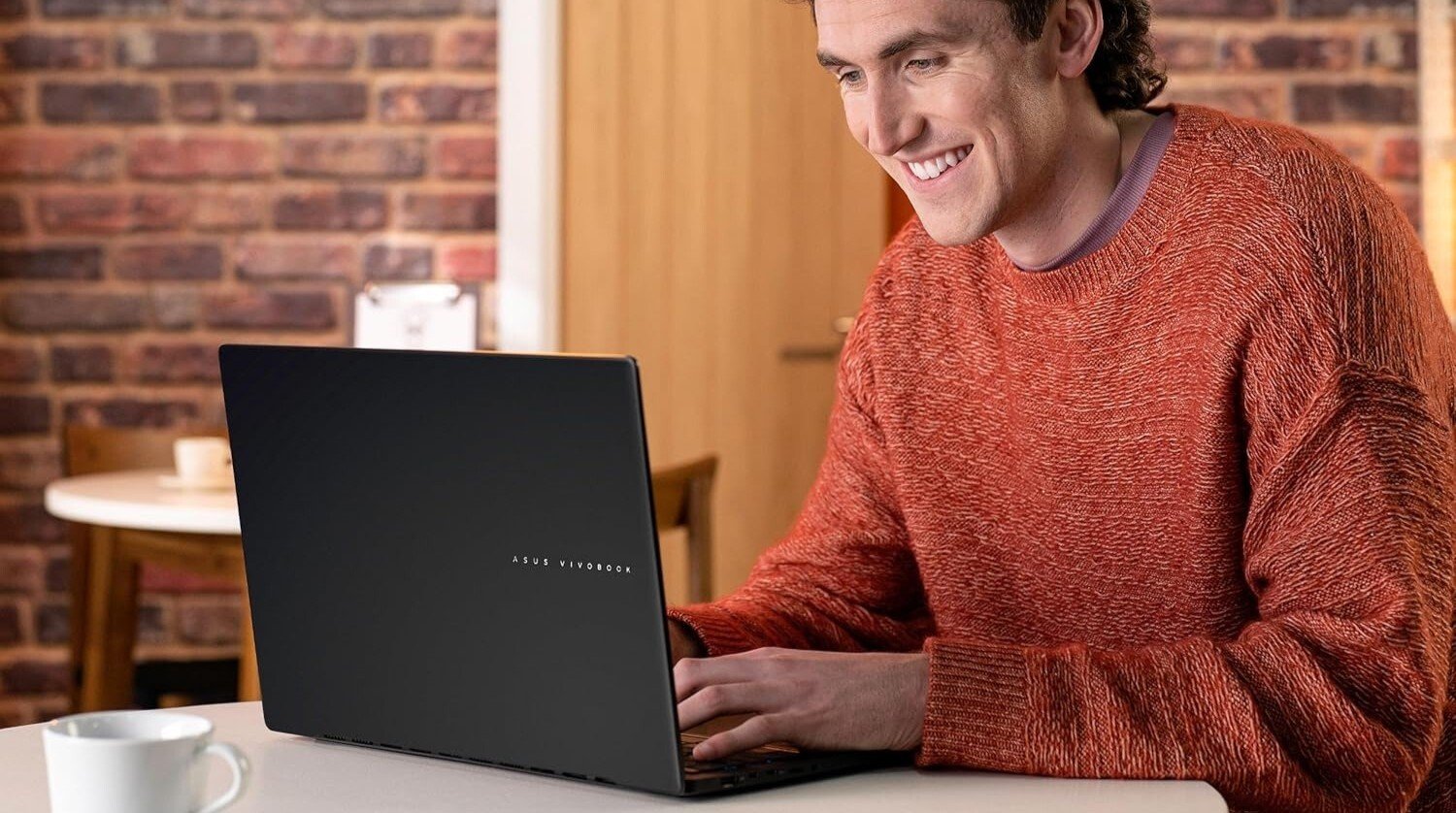a person sits at a table while looking at the Asus vivobook s16 laptop and smiling