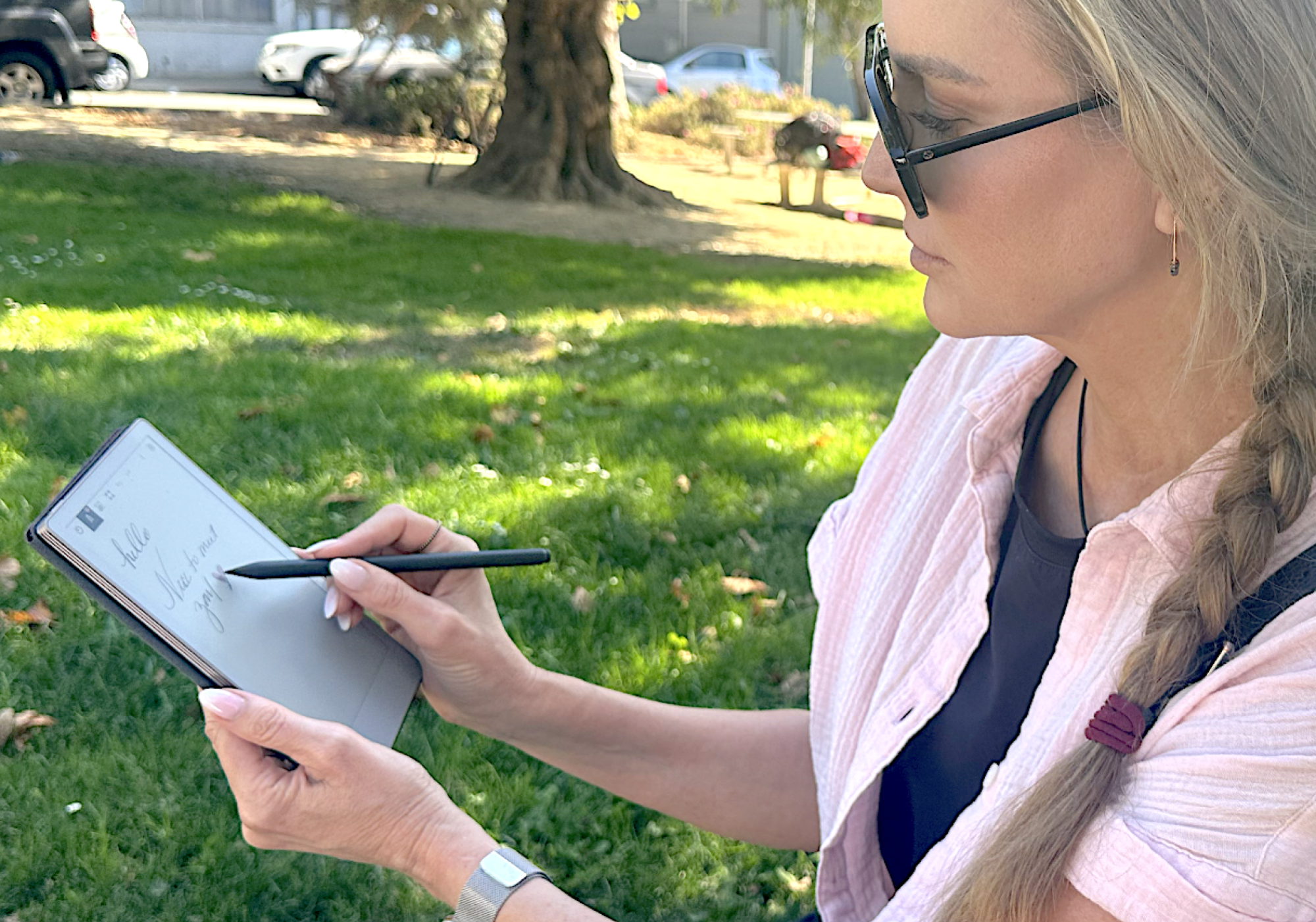 A woman in a park writes on a notebook-sized e-ink tablet held in one hand