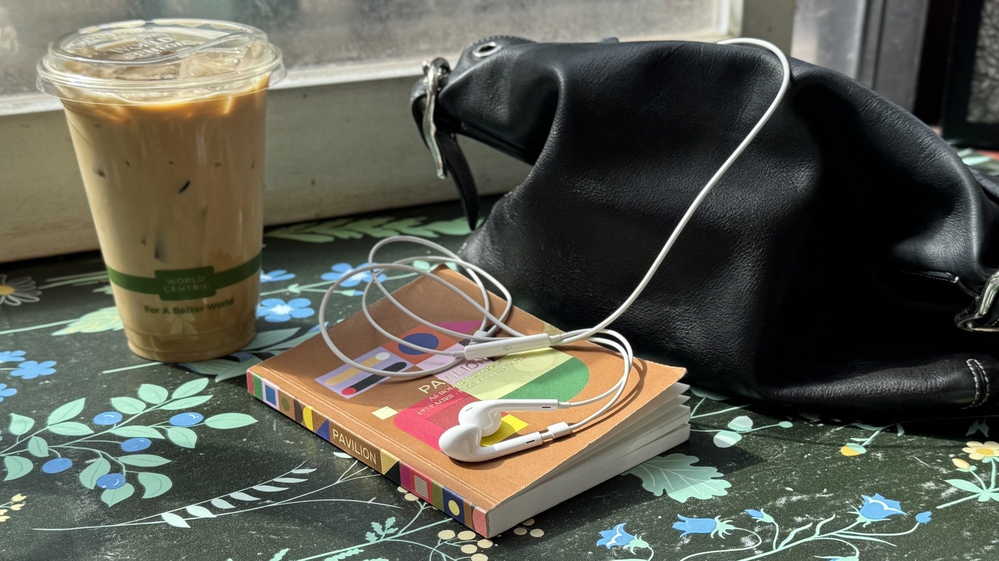 a purse with earpods sticking out of it next to an iced latte and a notebook on a counter