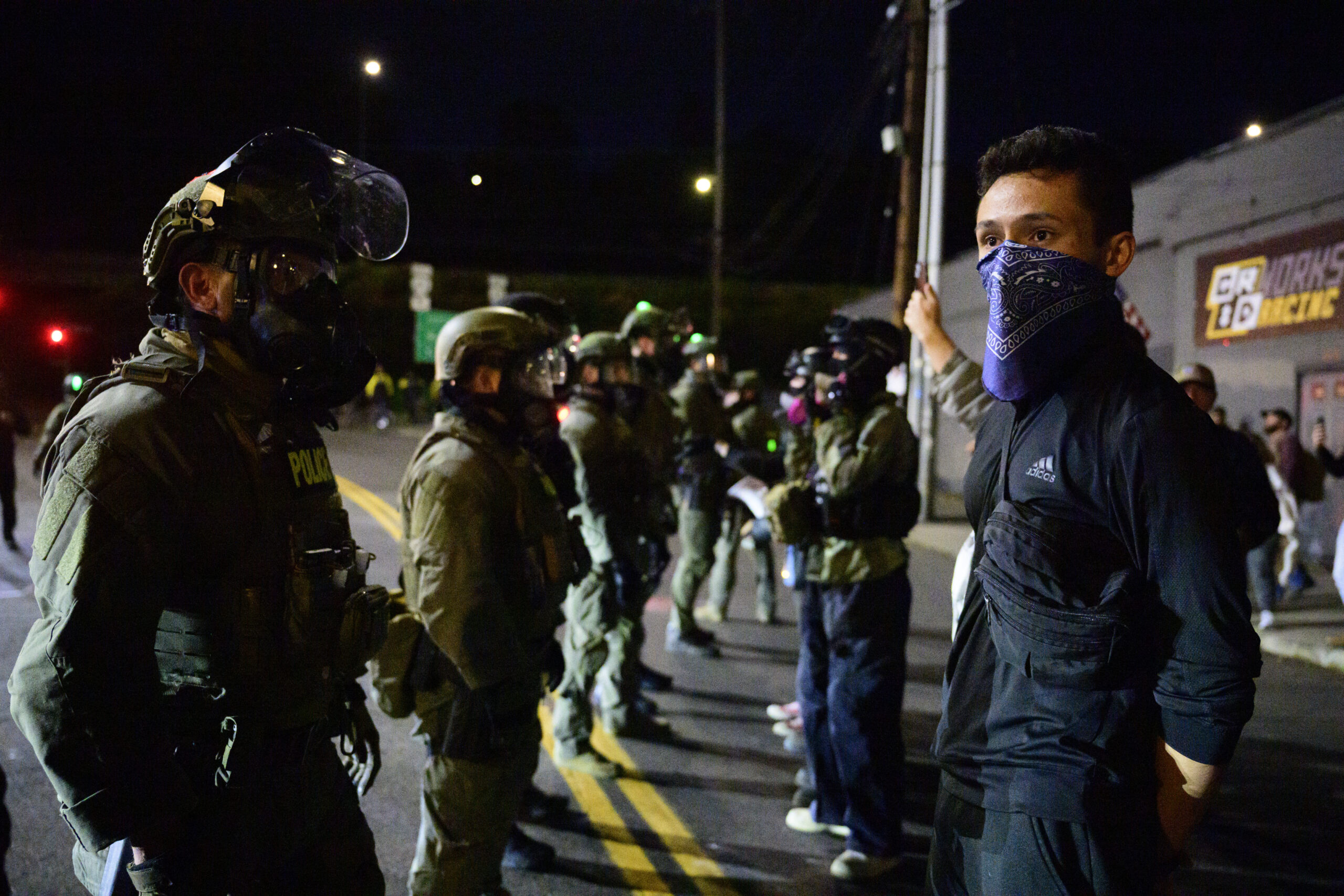 A line of masked and armed ICE officials stands facing a group of protesters.