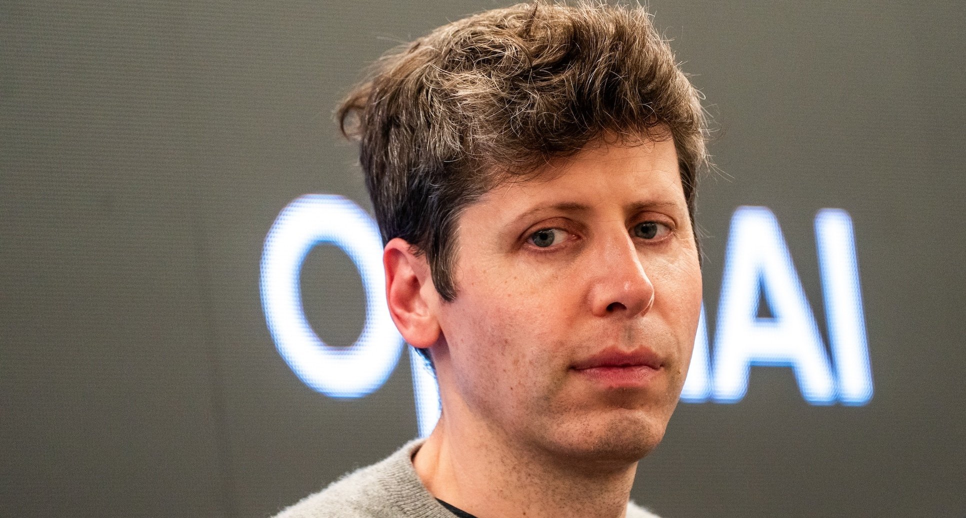 Sam Altman looking pensive before an OpenAI logo.