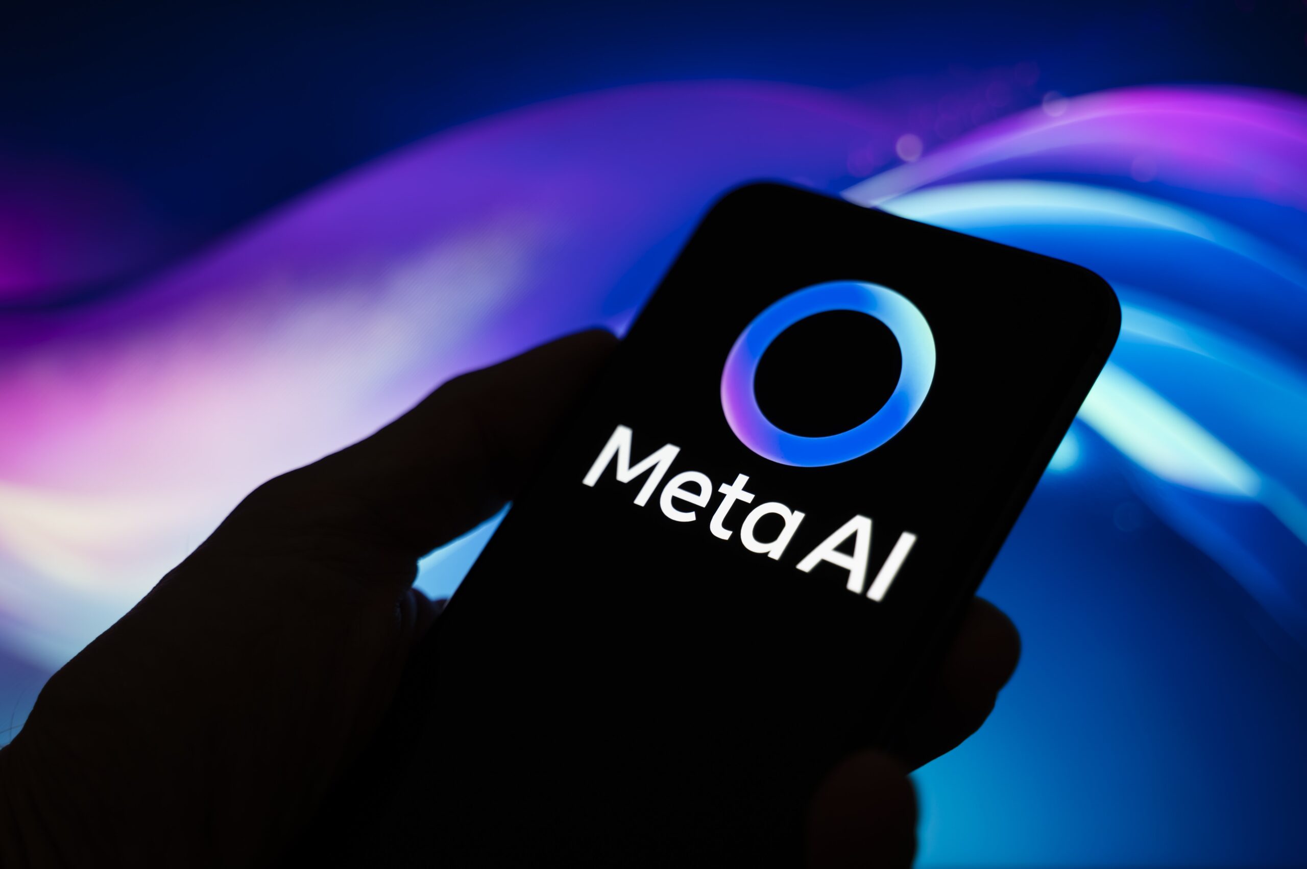 The Meta AI logo appears on a smartphone held in one hand with an abstract illustration of blue and purple colors in the background. Meta, the parent company of WhatsApp and Meta AI, makes the Artificial Intelligence assistant mandatory in its instant messaging app. 