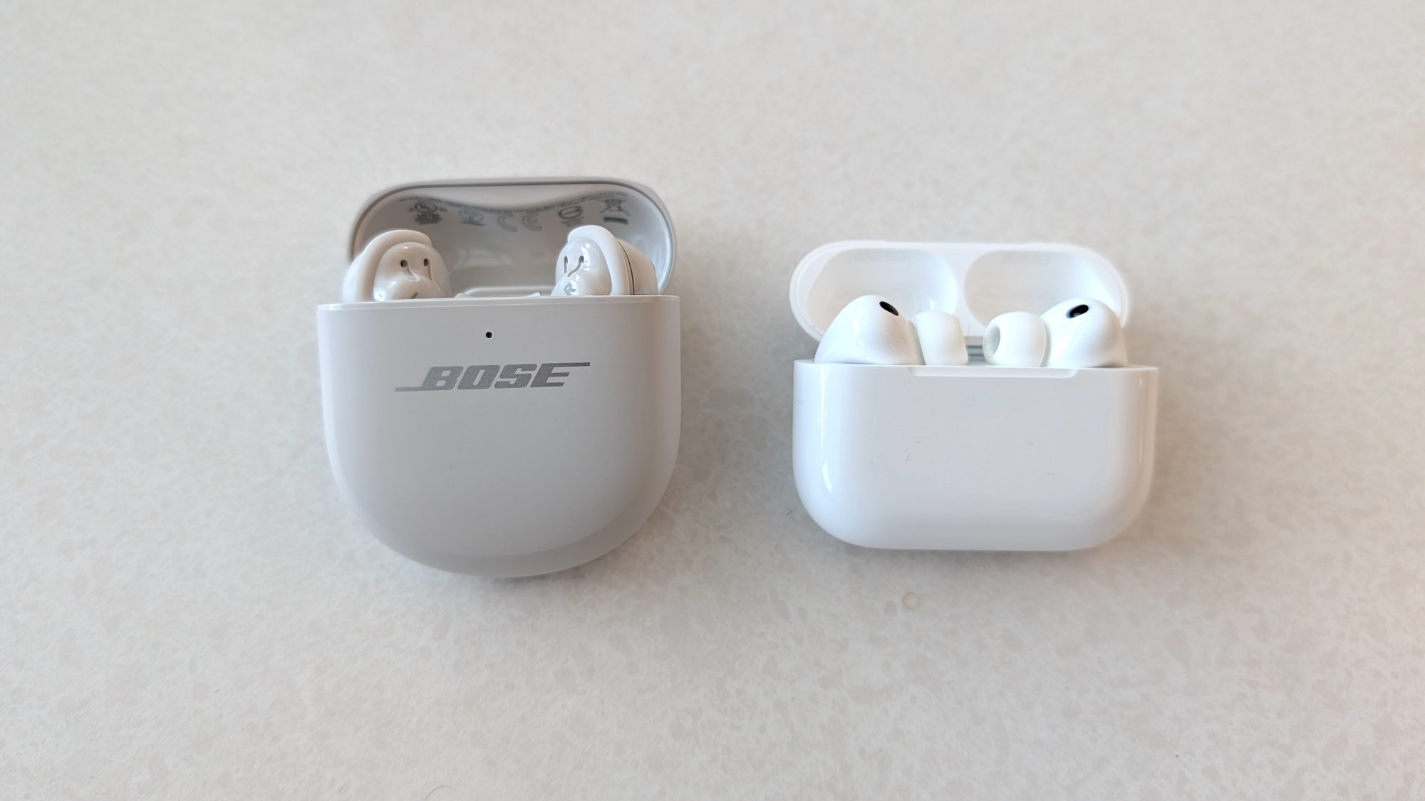 The Bose QuietComfort Ultra Earbuds (Gen 2) in their case, lying on a table next to the Apple AirPods Pro 3 in their case.