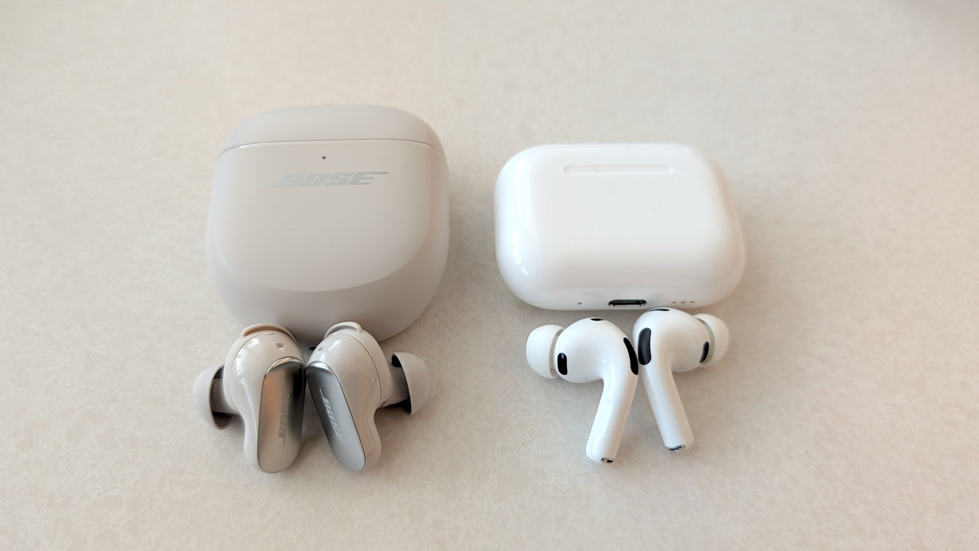 The Bose QuietComfort Ultra Earbuds (Gen 2) lying on a table next to the Apple AirPods Pro 3, both have their cases above them.