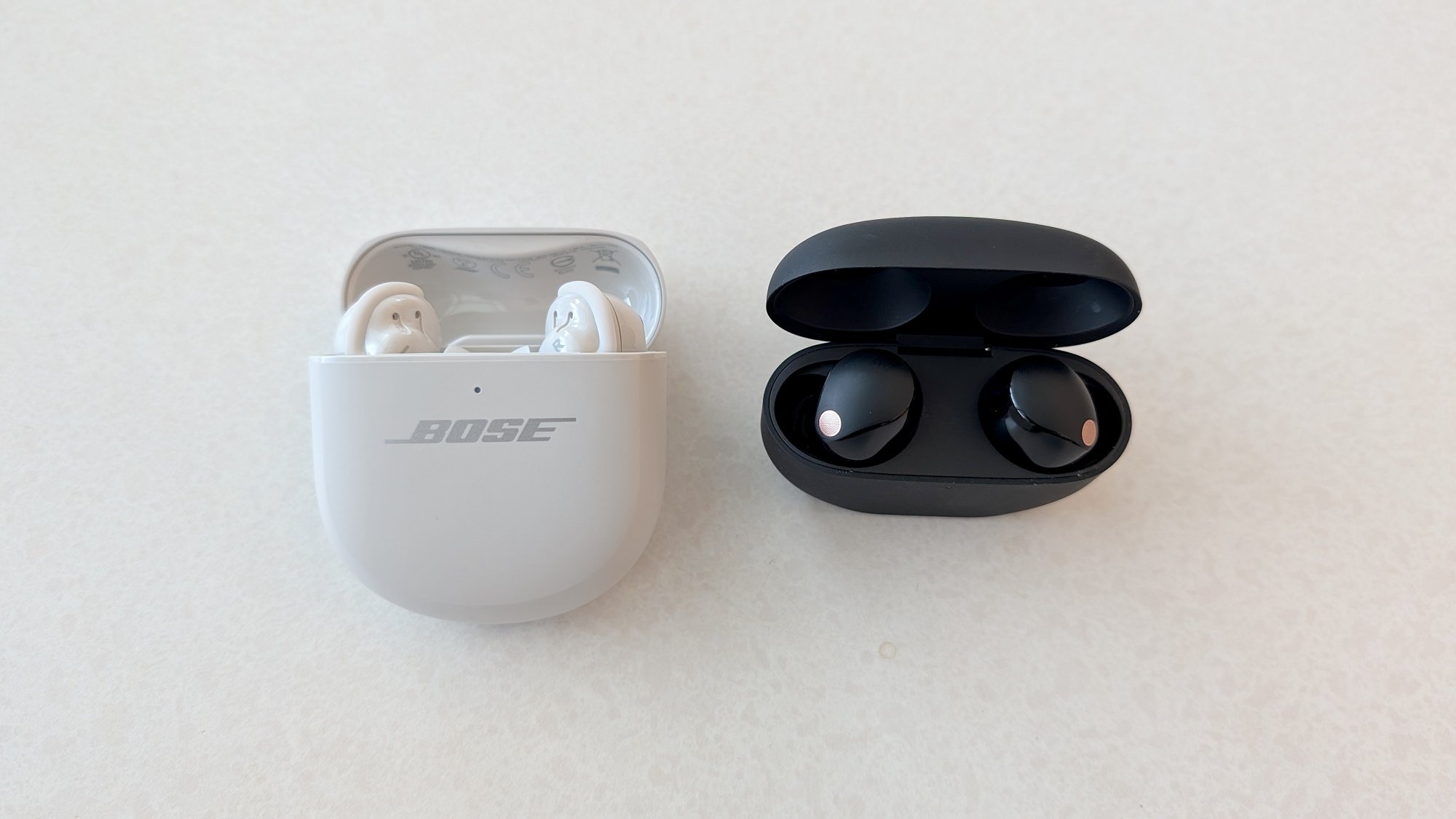The Bose QuietComfort Ultra Earbuds (Gen 2) in their case, lying on a table next to the Sony WF-1000xM5 earbuds in their case.