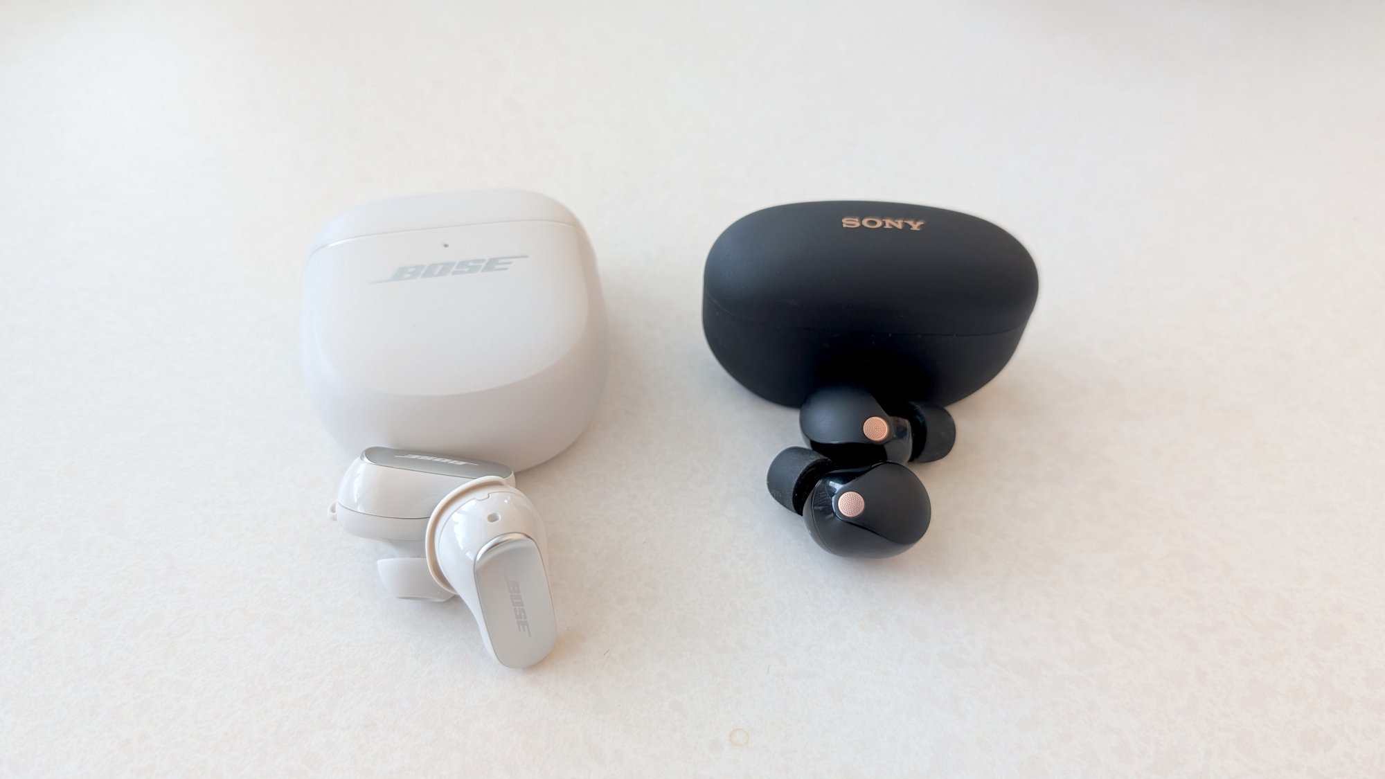 The Bose QuietComfort Ultra Earbuds (Gen 2) lying on a table next to the Sony WF-1000xM5 earbuds. Both have their cases above them.