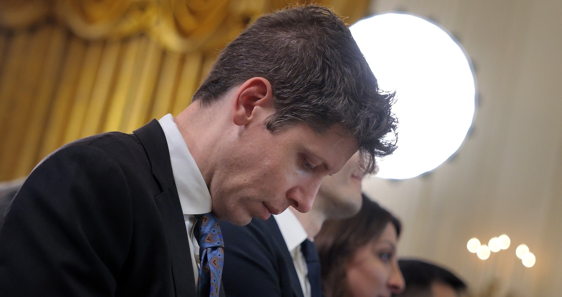 Sam Altman looks down in front of a gold curtain and a round light