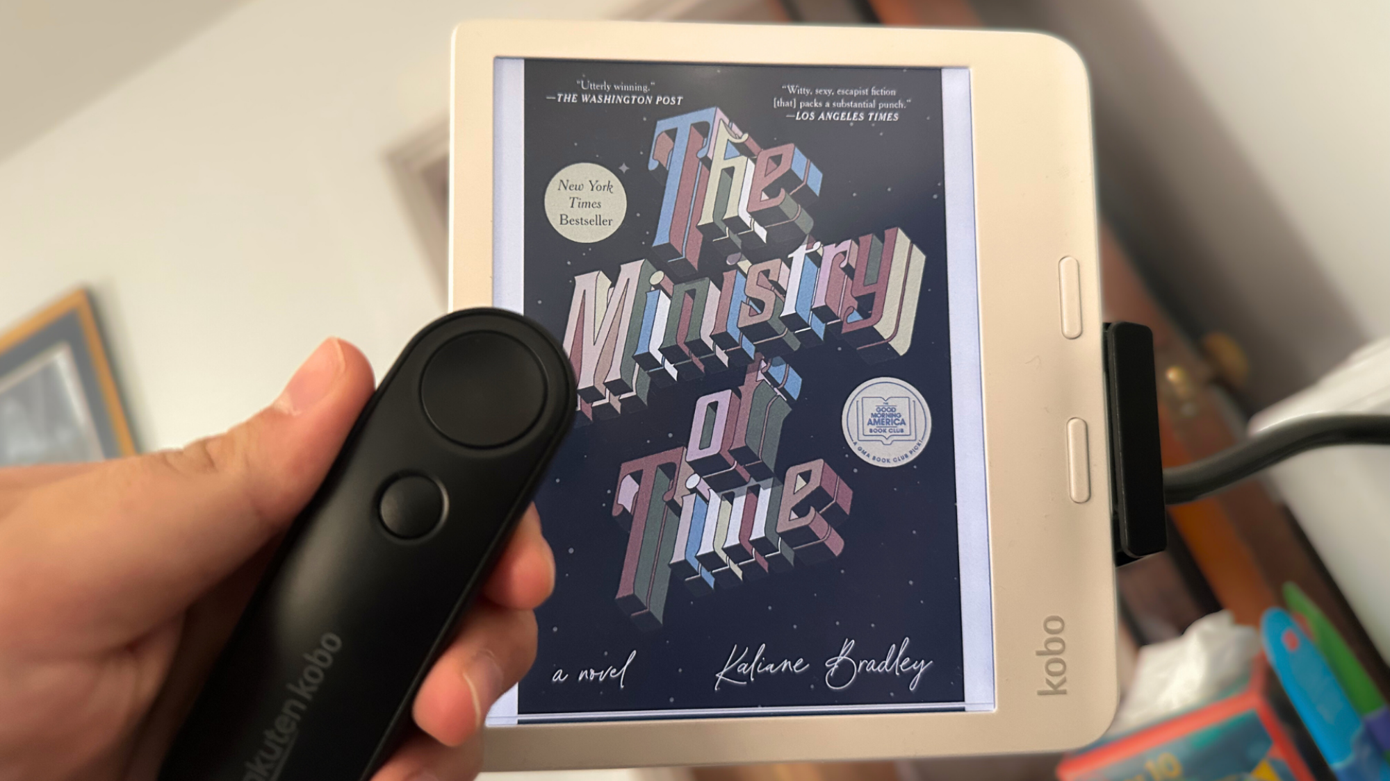 A Kobo Libra Colour in a stand with a Kobo Remote being held in someone's hand.