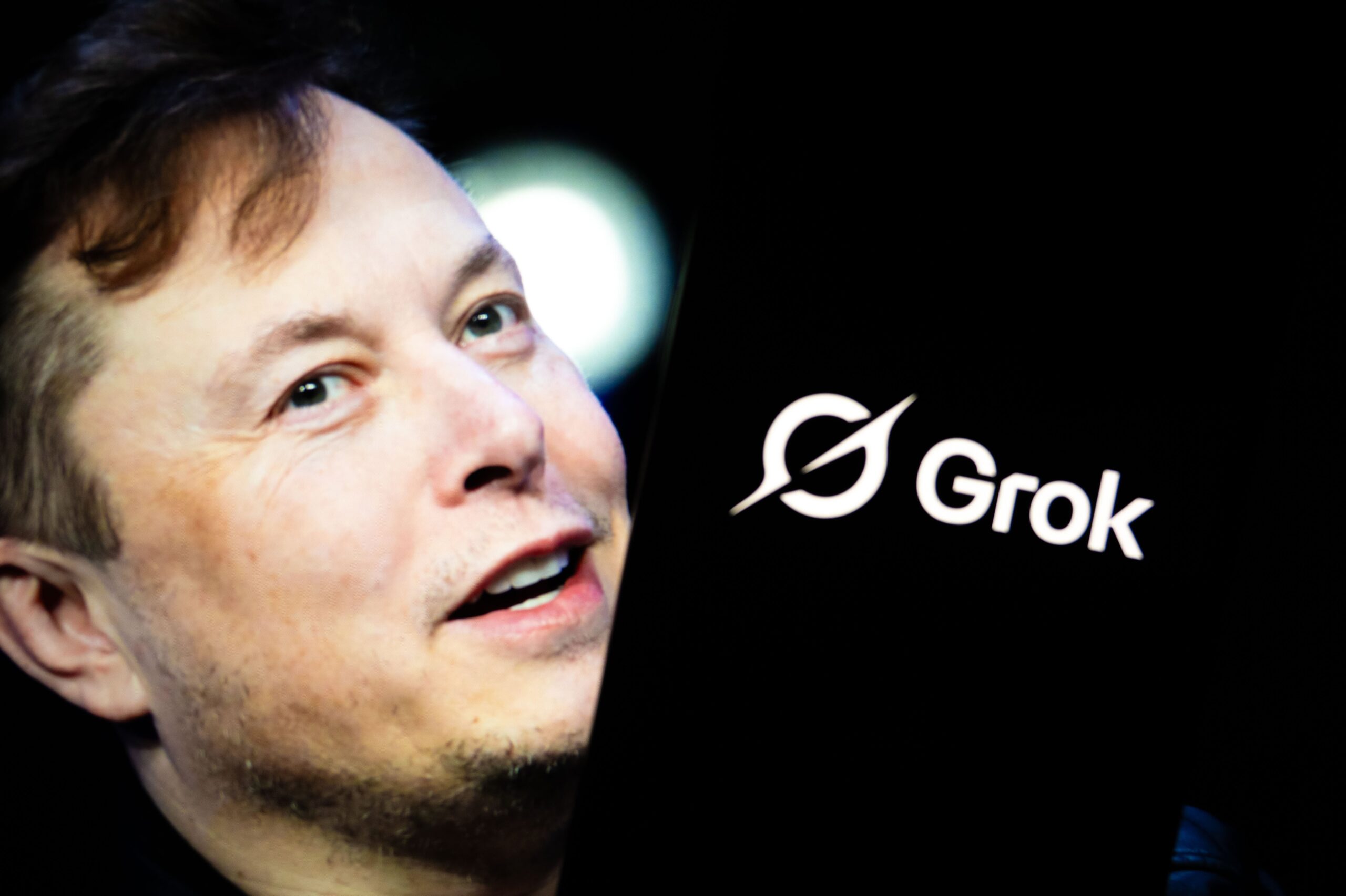 'Grok' logo displayed on a smartphone screen in front of a picture of Elon Musk