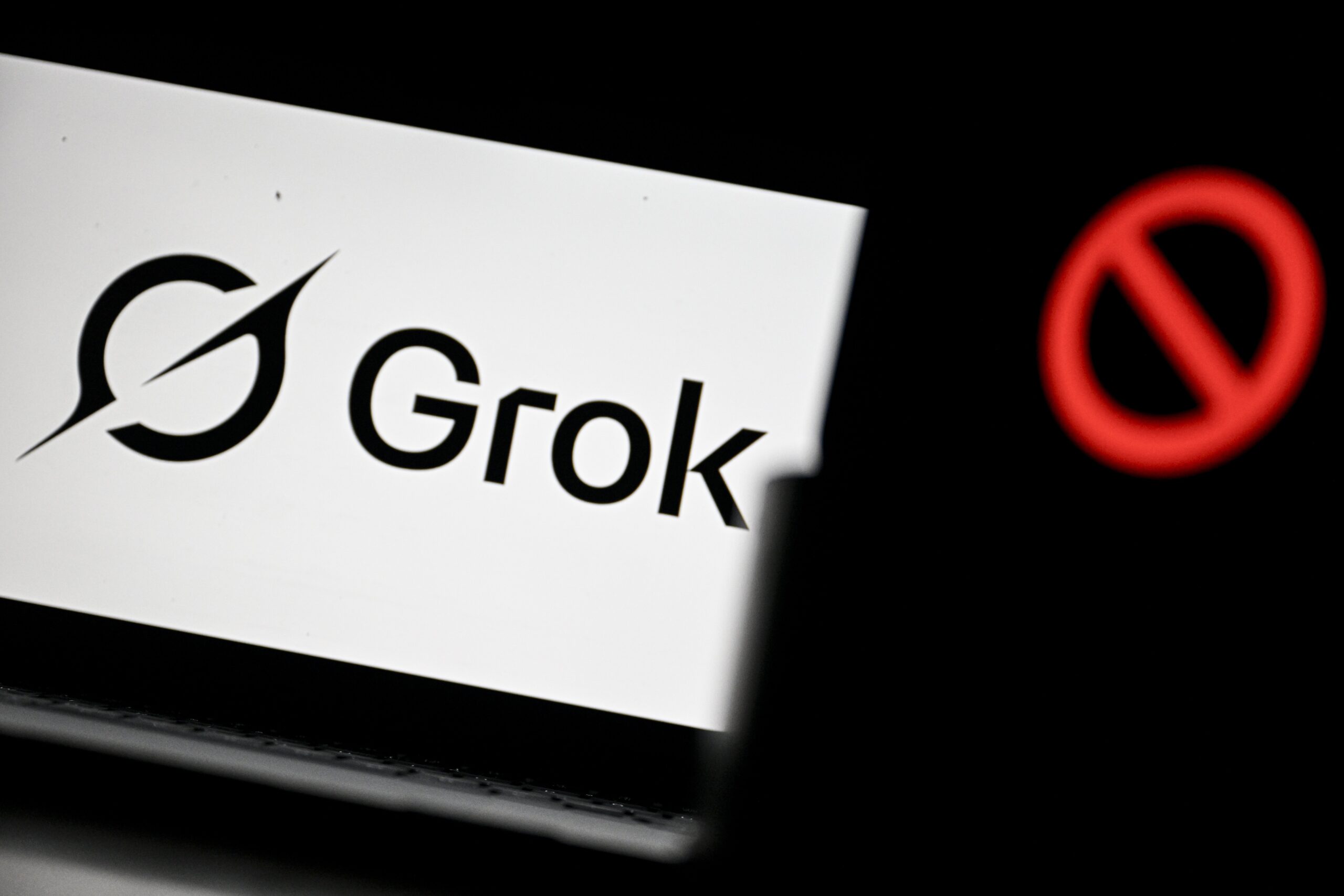 In the background: a laptop screen showing the Grok logo. In the foreground is a large red no symbol on a phone. 