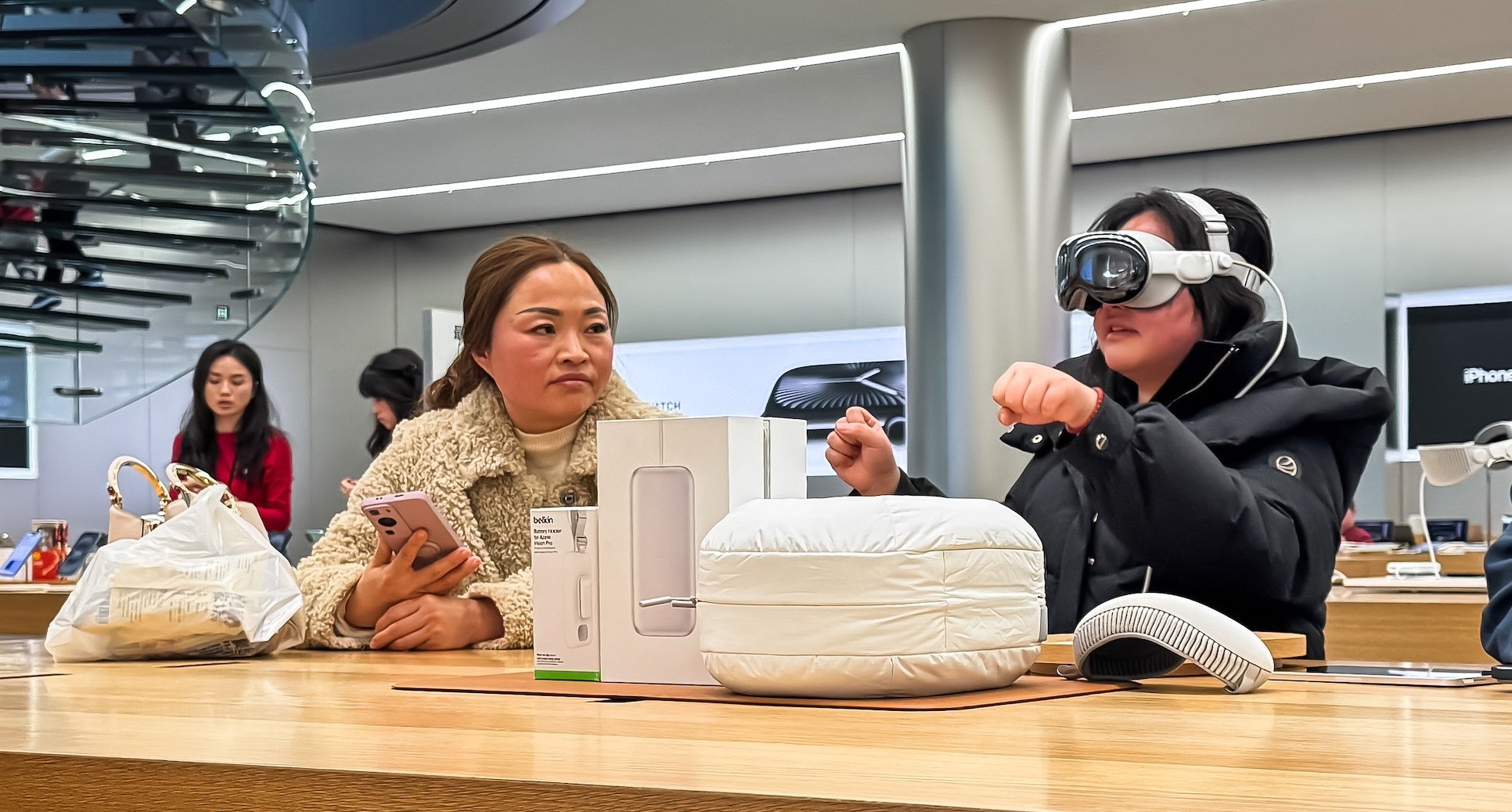 A woman holding an iPhone in an Apple store looks on as another woman wears the Vision Pro.