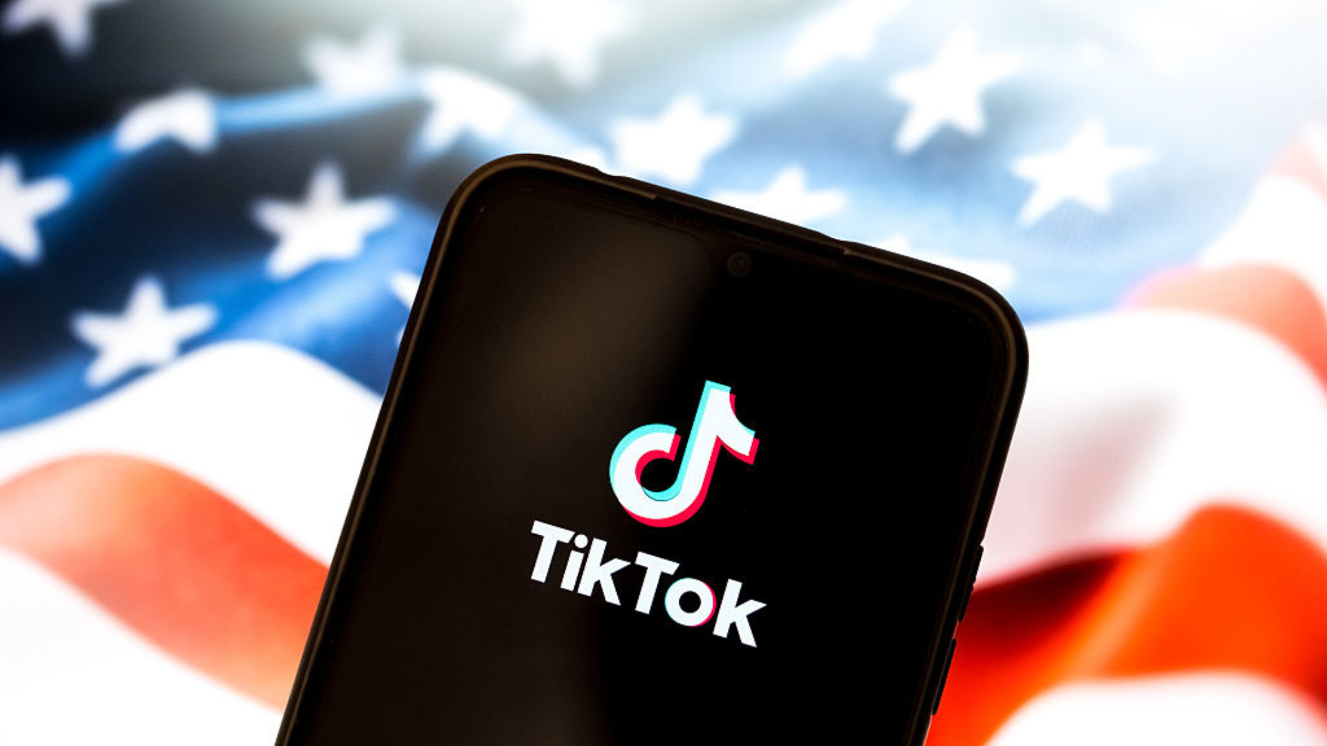 The TikTok logo appears on a smartphone screen, with the American flag on a computer screen in the background.