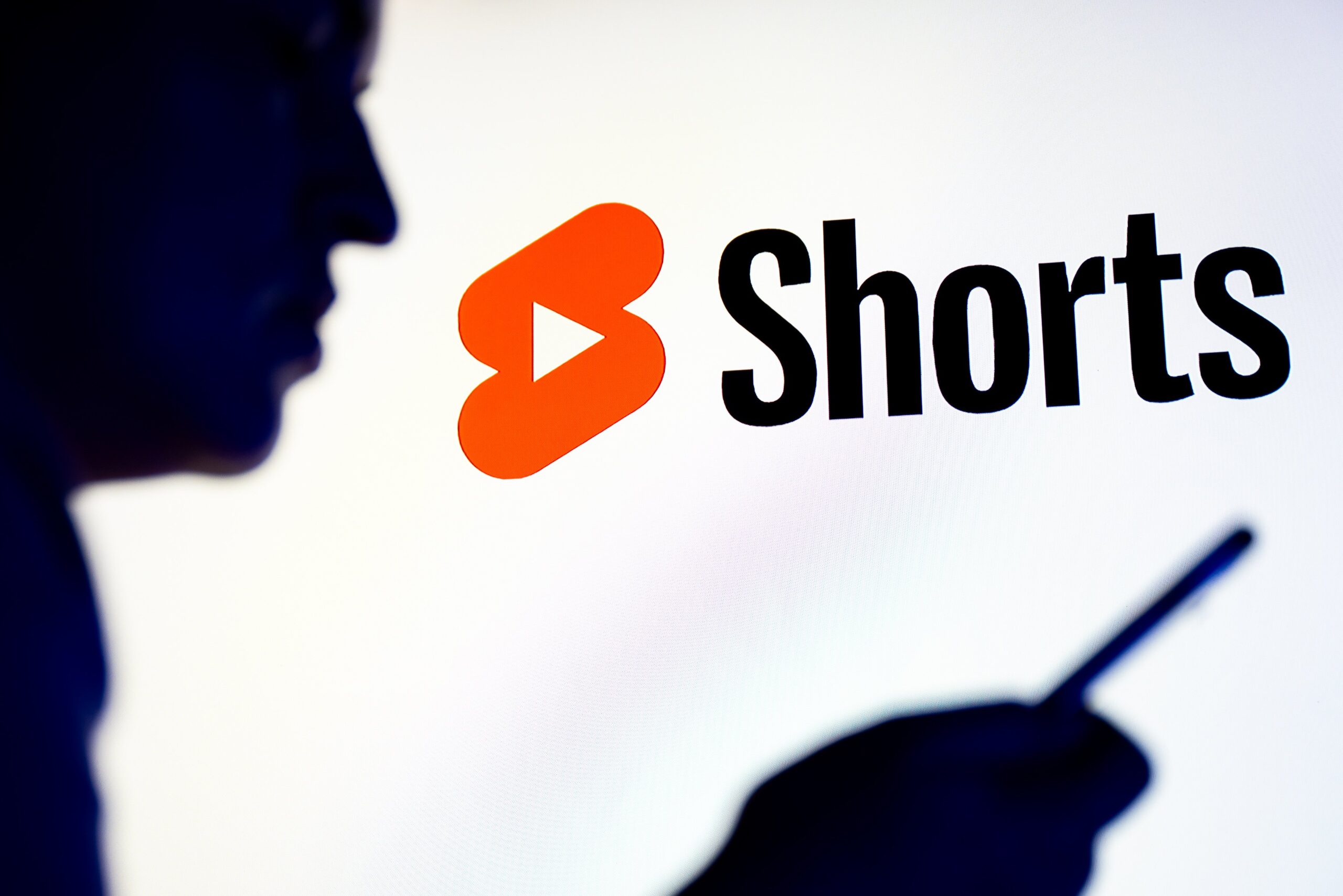 A person stands in profile staring at a phone. A large YouTube Shorts logo glows behind them. 