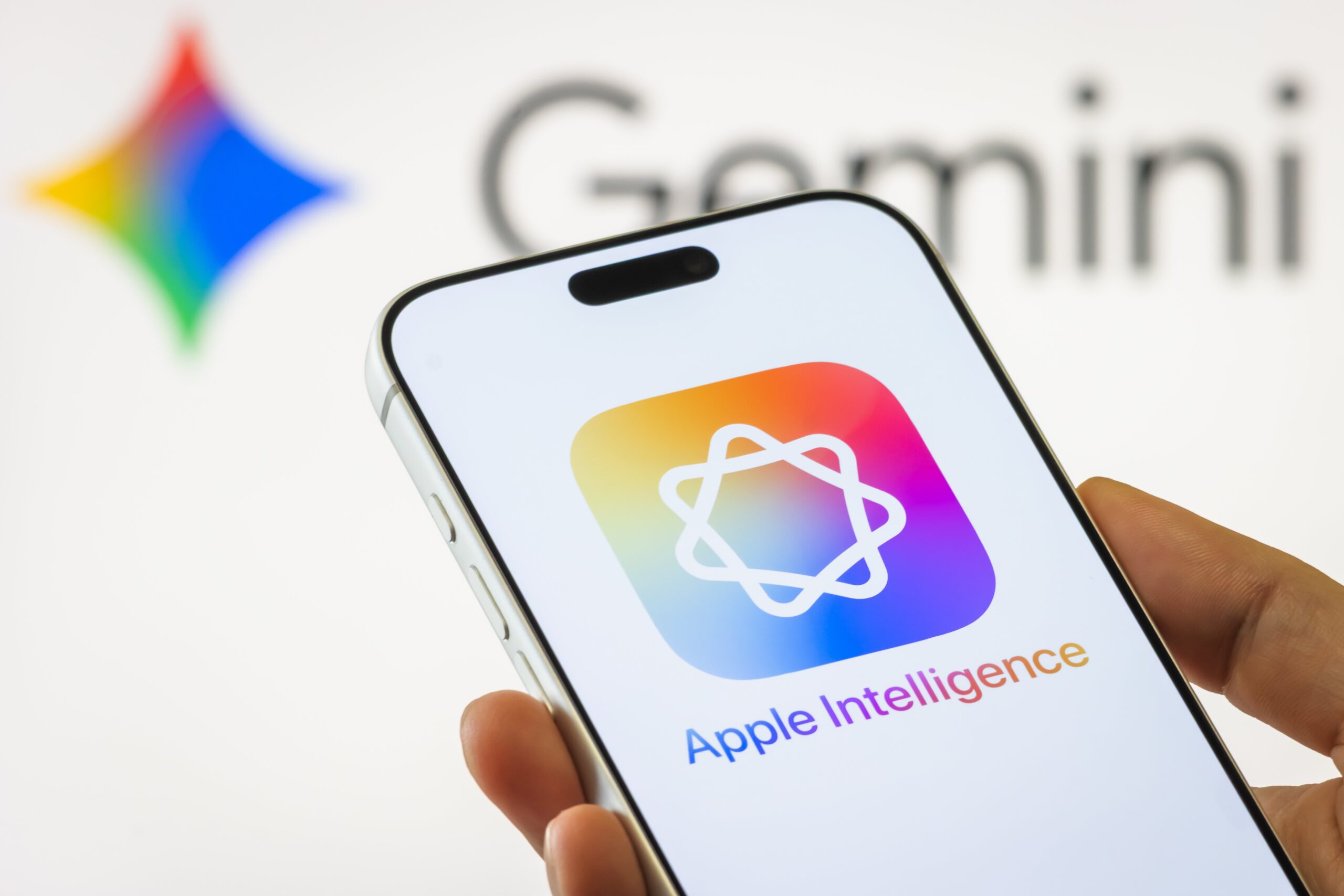 A hand holding a phone with the Apple Intelligence logo in front of a screen with the Gemini logo.