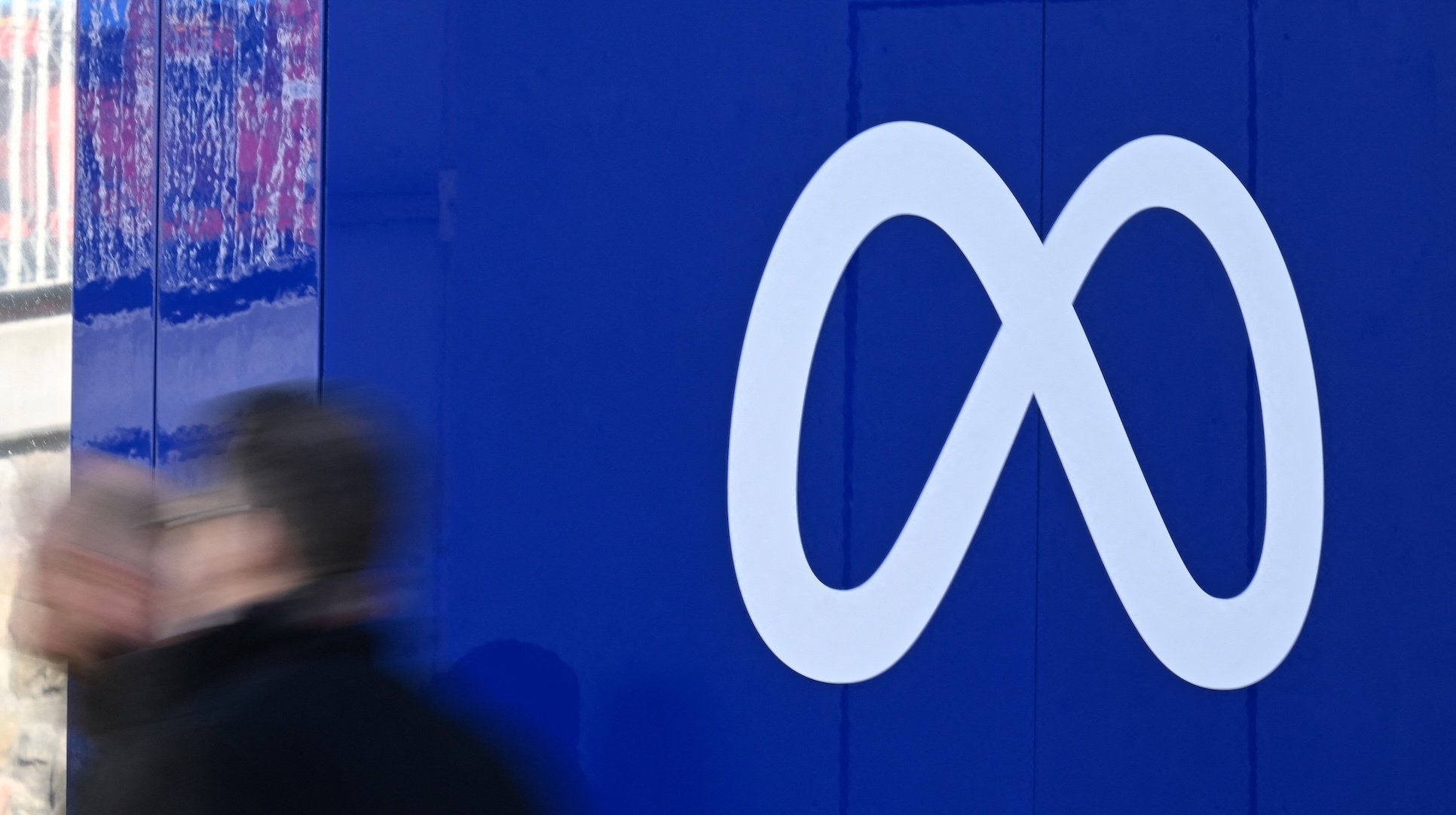 The Meta logo, white on blue, with blurred figures passing by. 