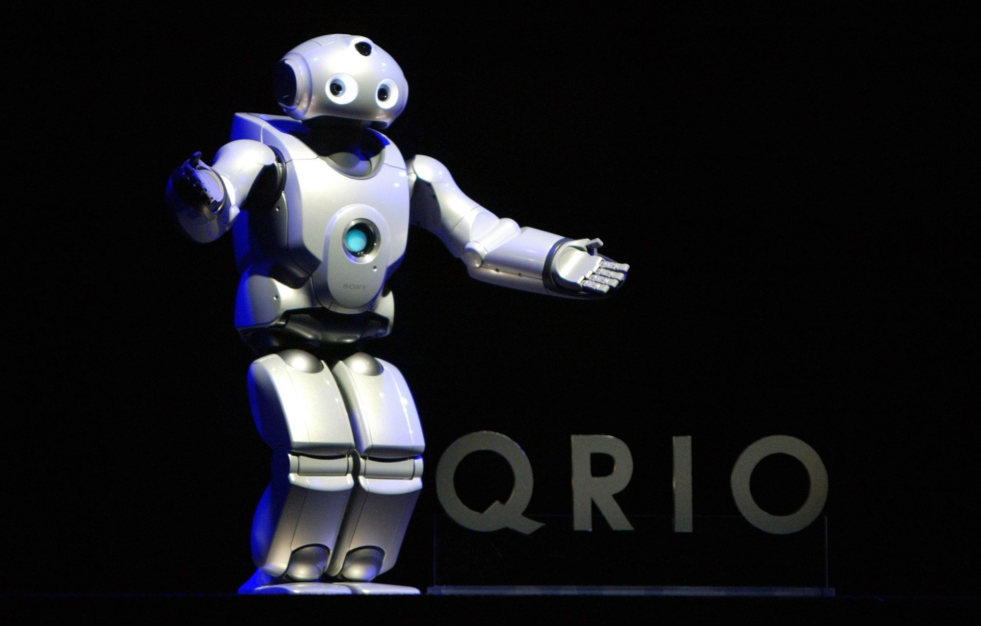 A humanoid robot with green chest light waves on stage in front of a QRIO sign