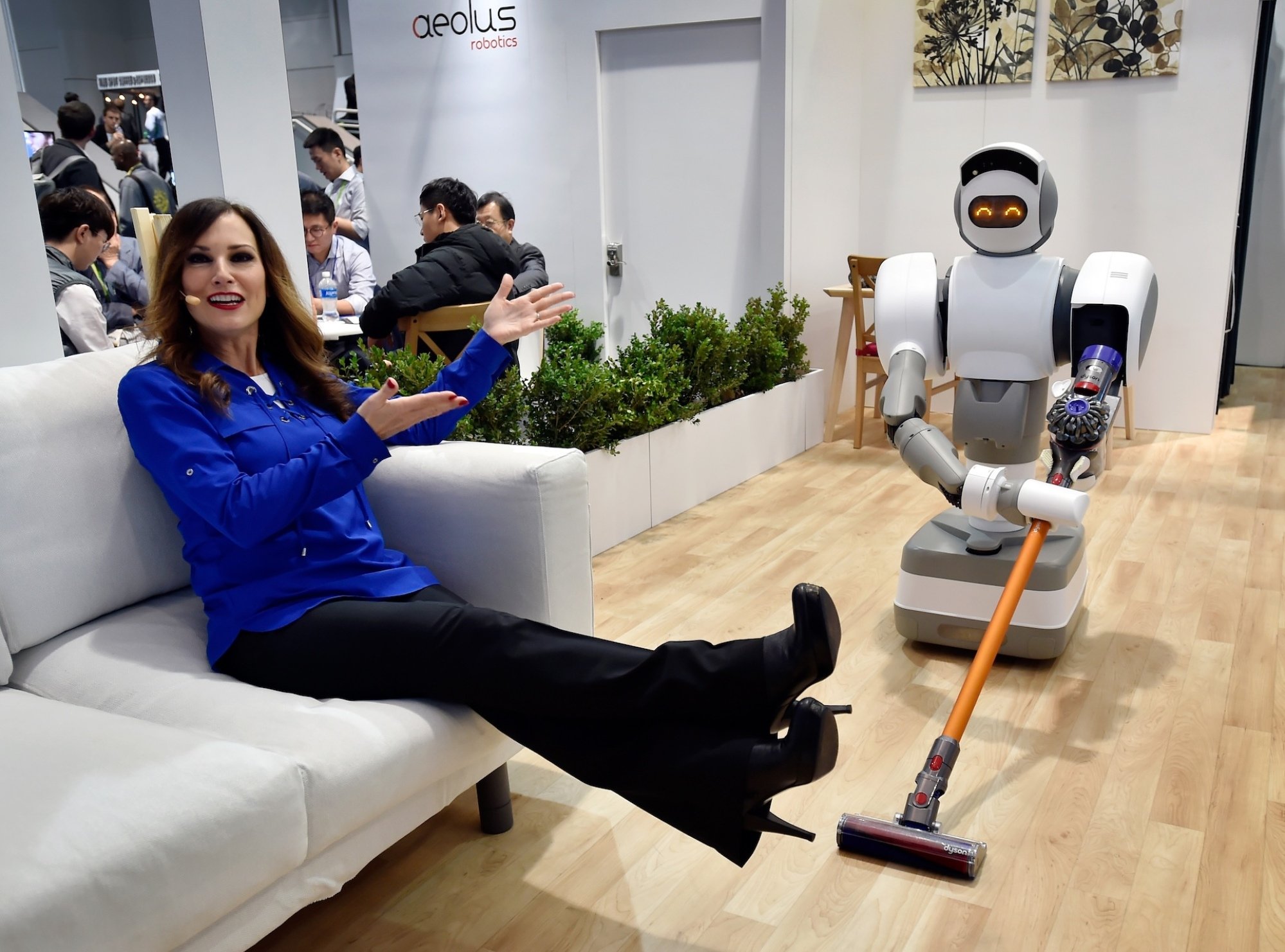 A woman on a couch shows off a robot pushing a vacuum cleaner
