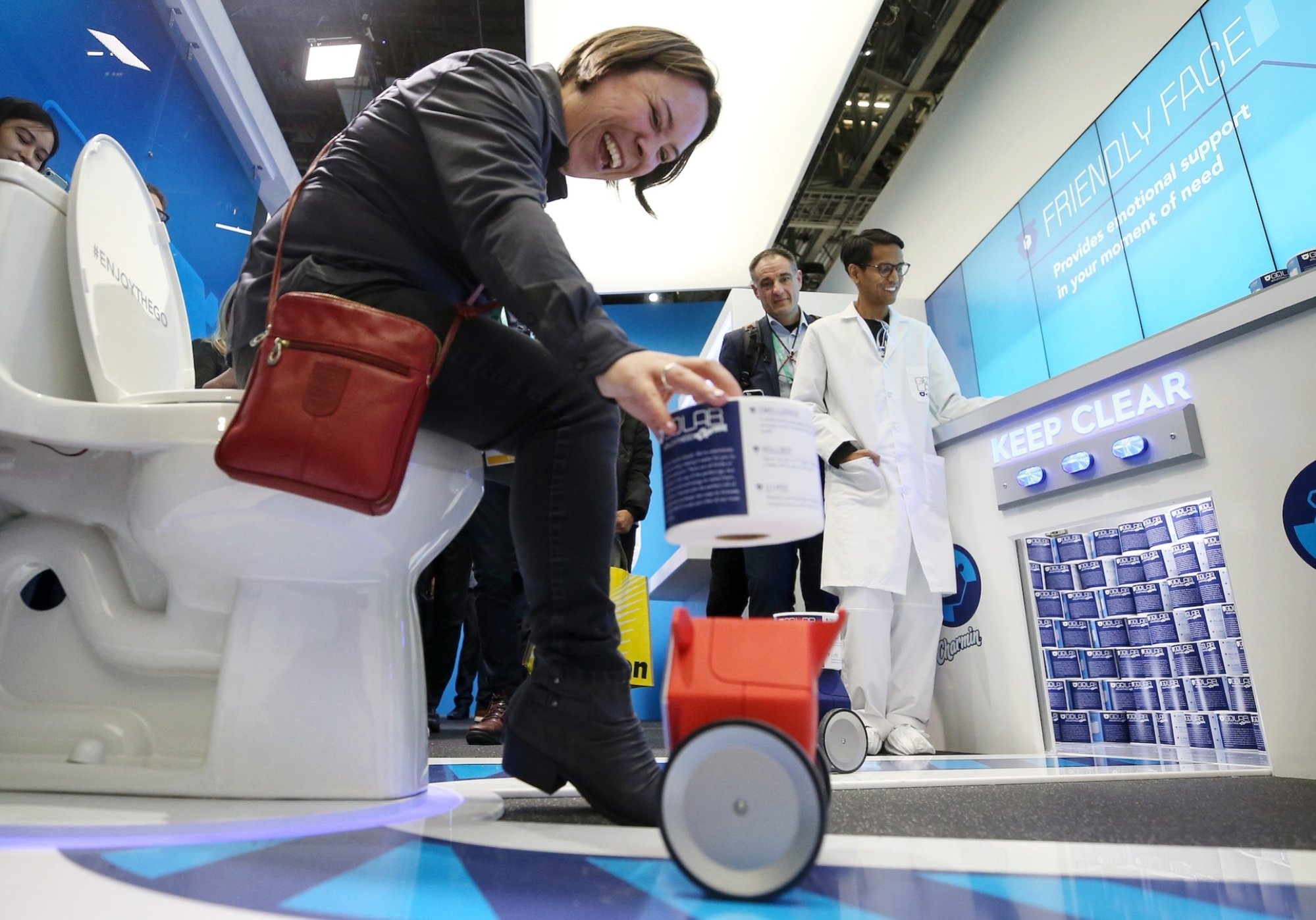 A woman on a toilet receives a roll of paper from a small wheeled robot.
