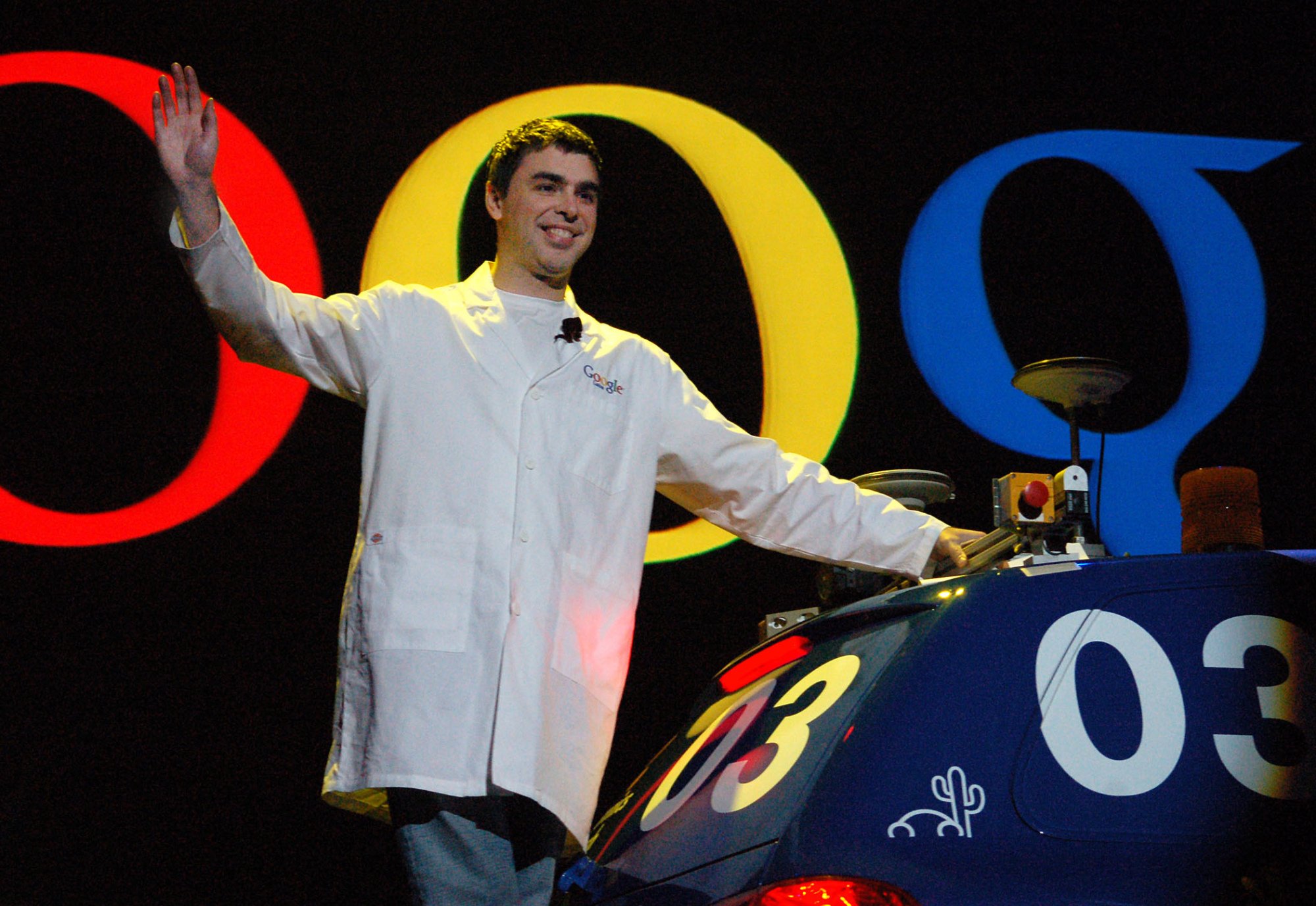 Larry Page in a white coat riding a robot car.