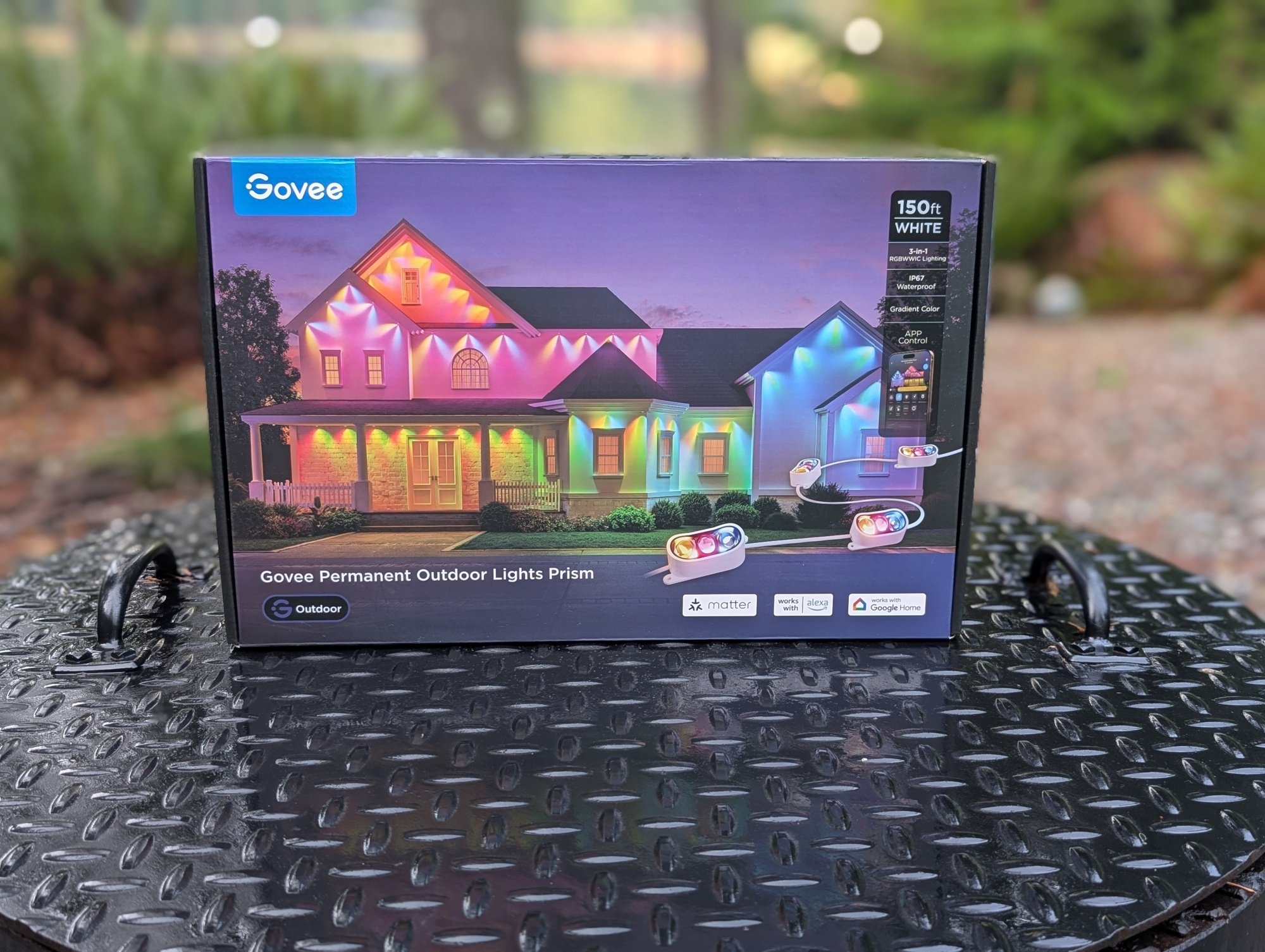 Govee Permanent Outdoor Lights Prism in their box sitting outside with a forest scene behind the box.