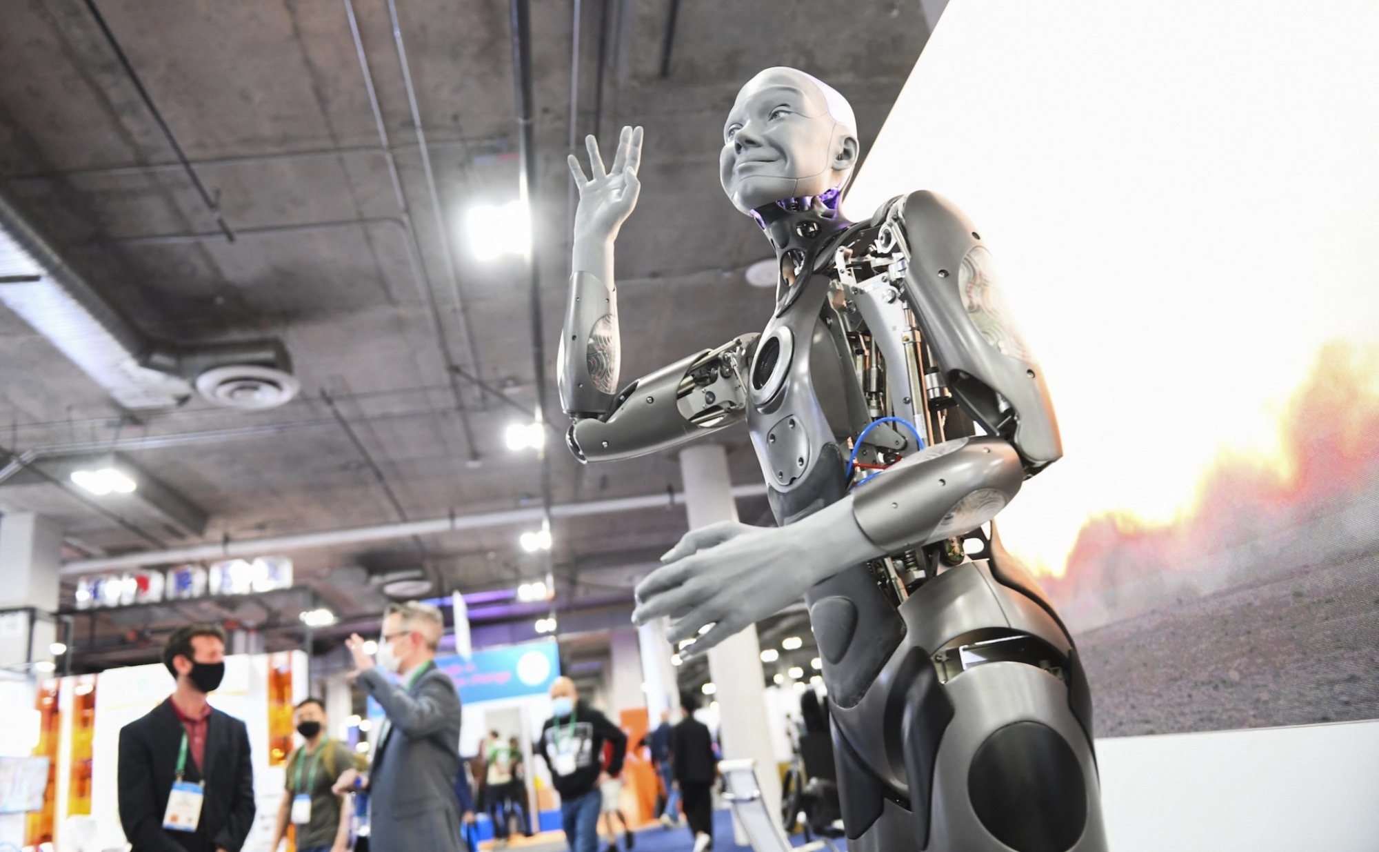 A silver robot with a very realistic face waves at attendees with masks on.