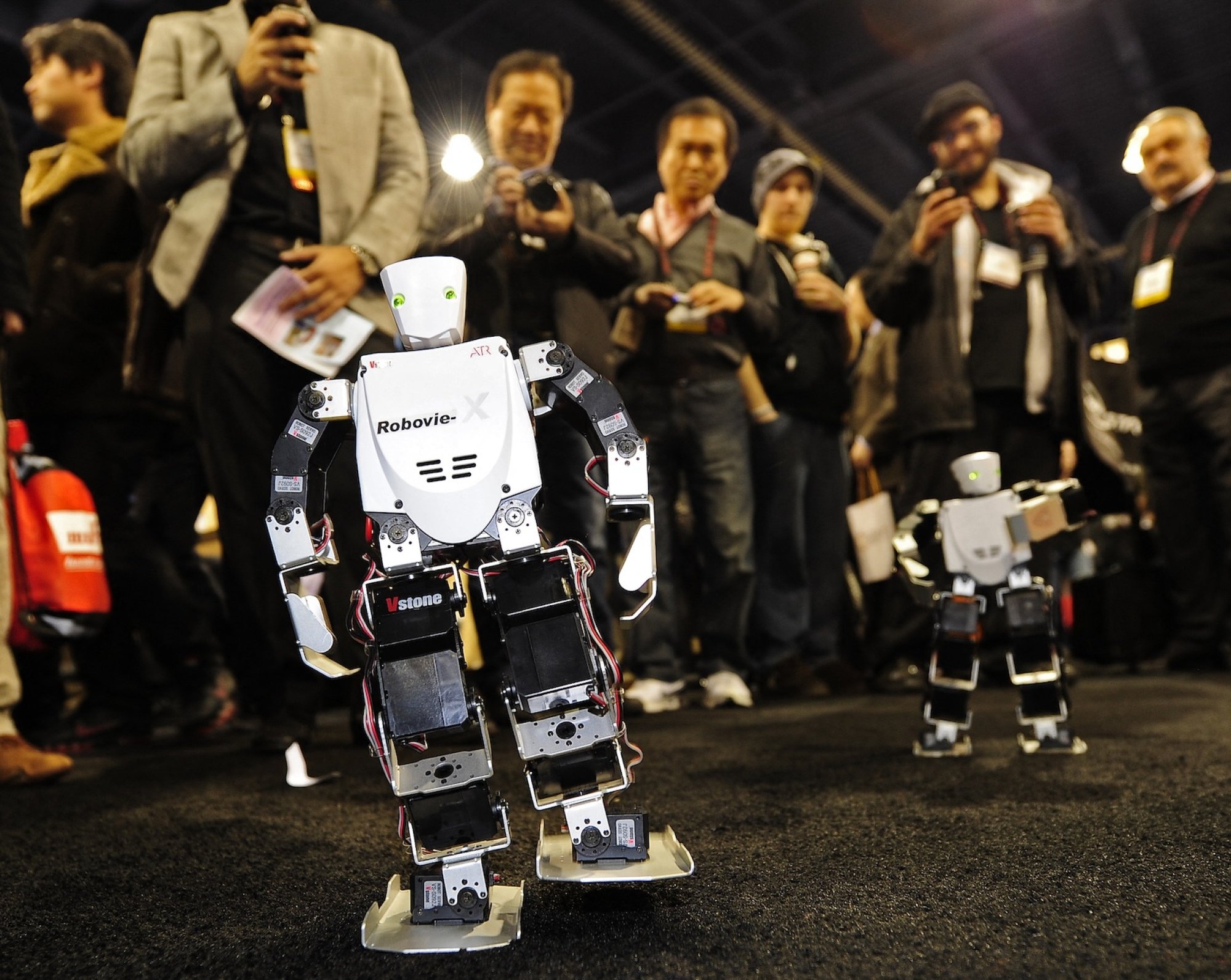 Two small robots walk past attendees with cameras