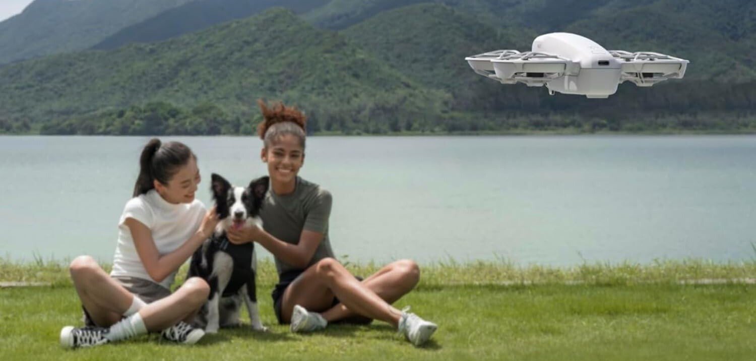 two people sit on a grass field with a dog while the DJI Neo drone flies in front of them