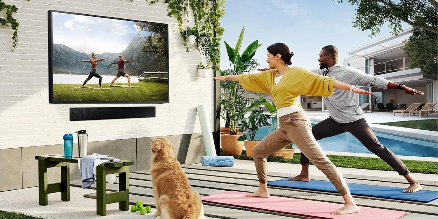 the Samsung The Terrace TV mounted outside under an awning while the TV shows a yoga video. Two people and a dog are watching the yoga video.
