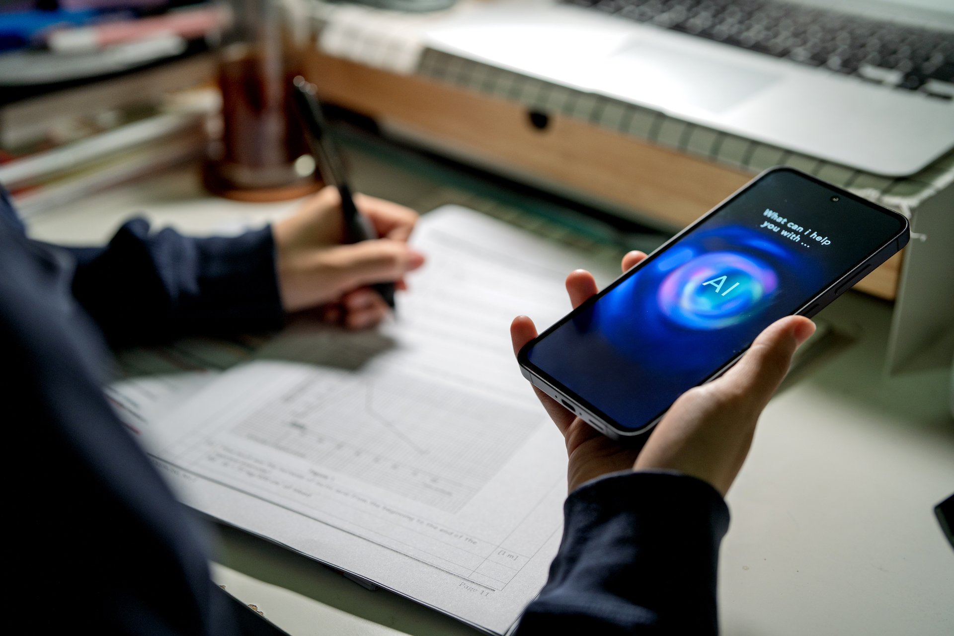 A teen holds a phone in their hand and consults an AI for help while writing in a notebook. 