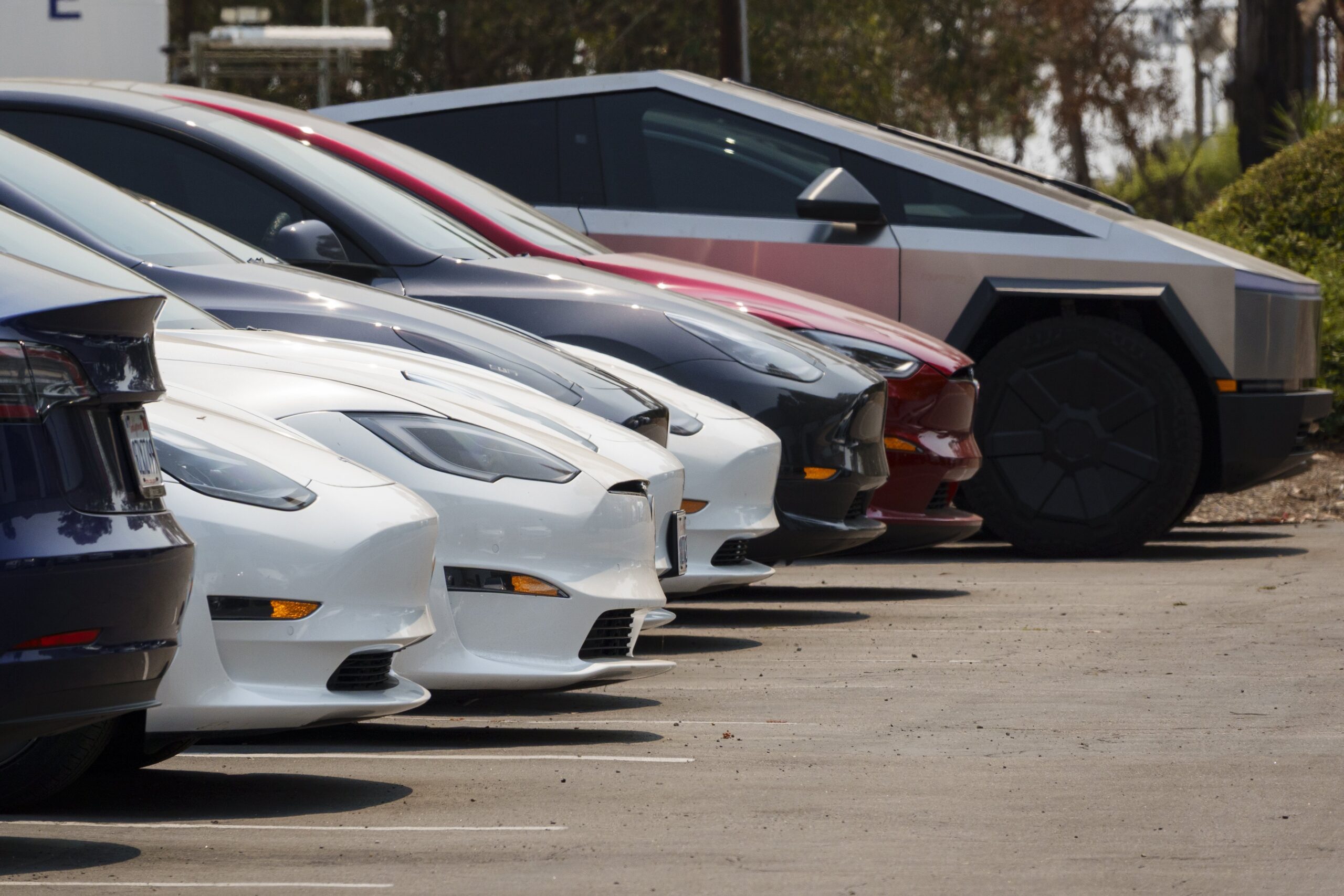 A row of Tesla EVs and a cybertruck in a sunny parking lot.
