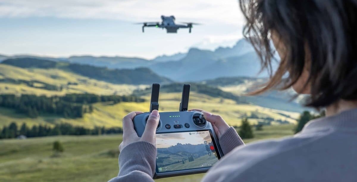 a person flies the DJI Mini 5 Pro drone in a green mountainous area. the image views the drone and the DJI RC from over the shoulder of the operator