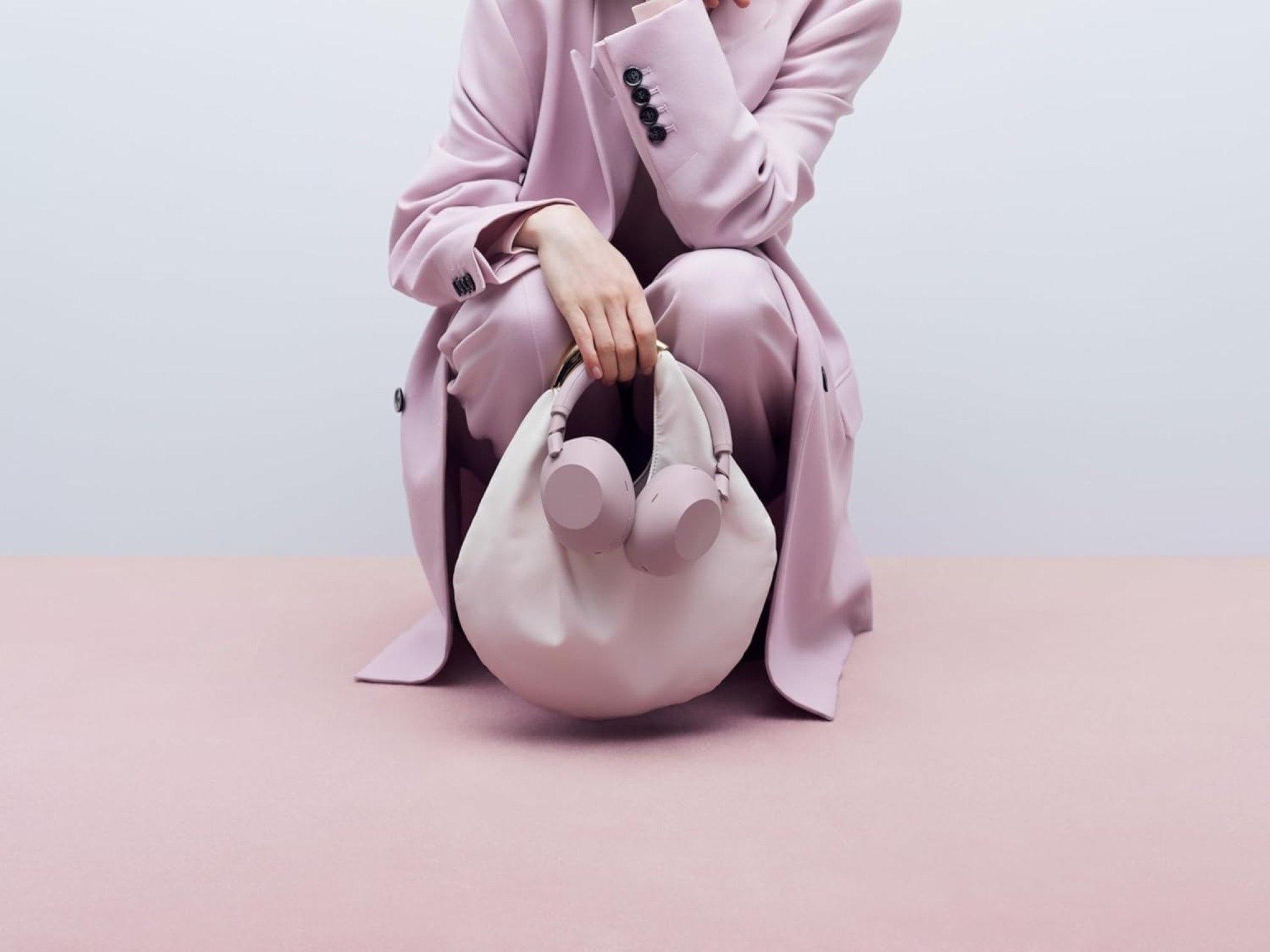 a person crouches down while wearing all pink and holding the sony XM6 headphones in sand pink.