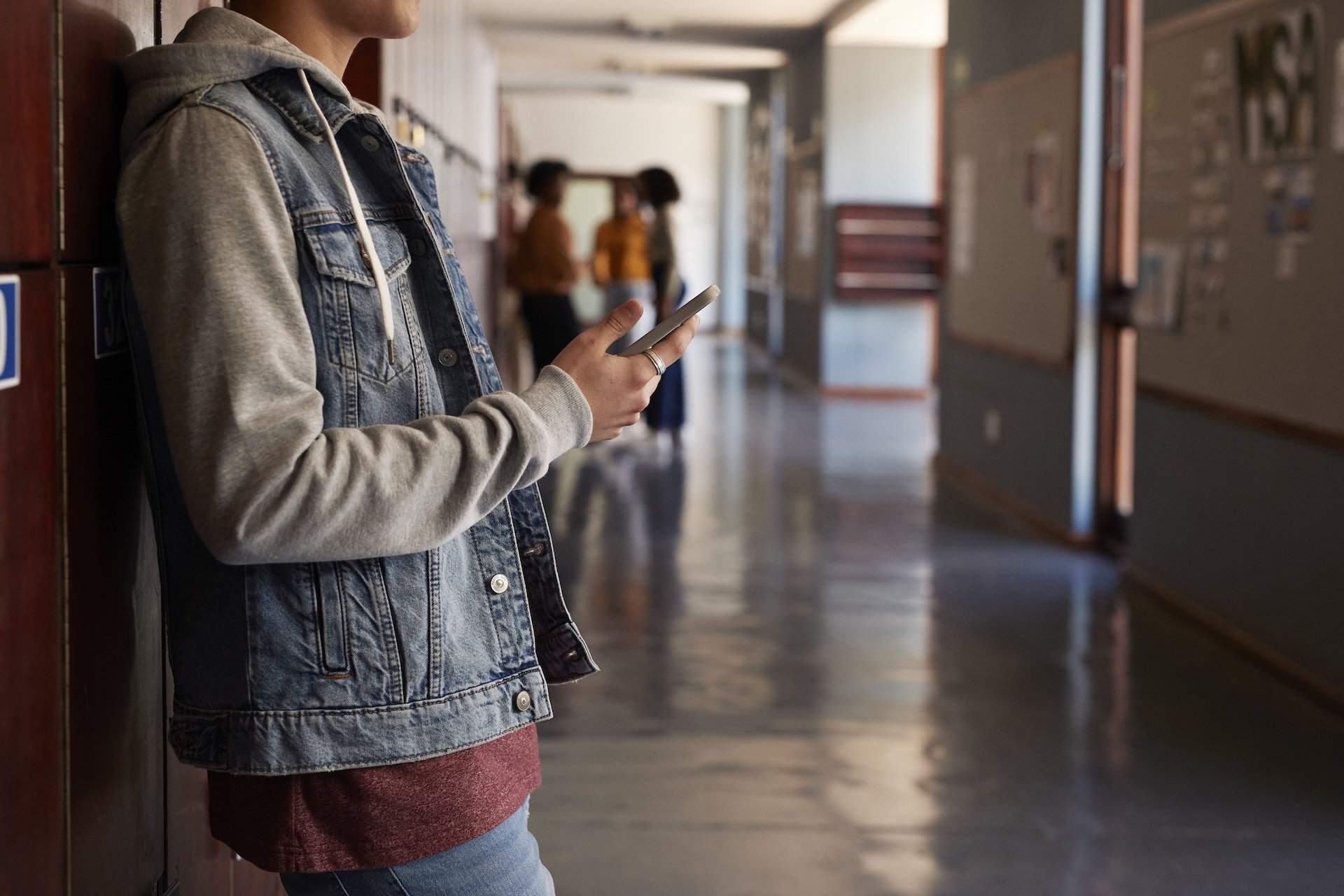 Teen boy stands in school hallway holding phone in his hand. 
