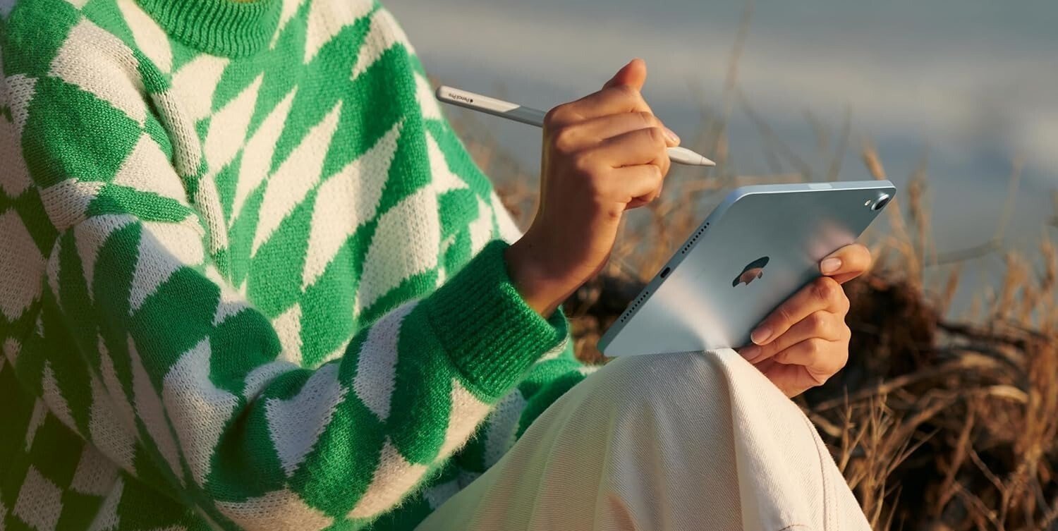 a person sits on the beach while holding the ipad mini and using an apple pencil with the tablet
