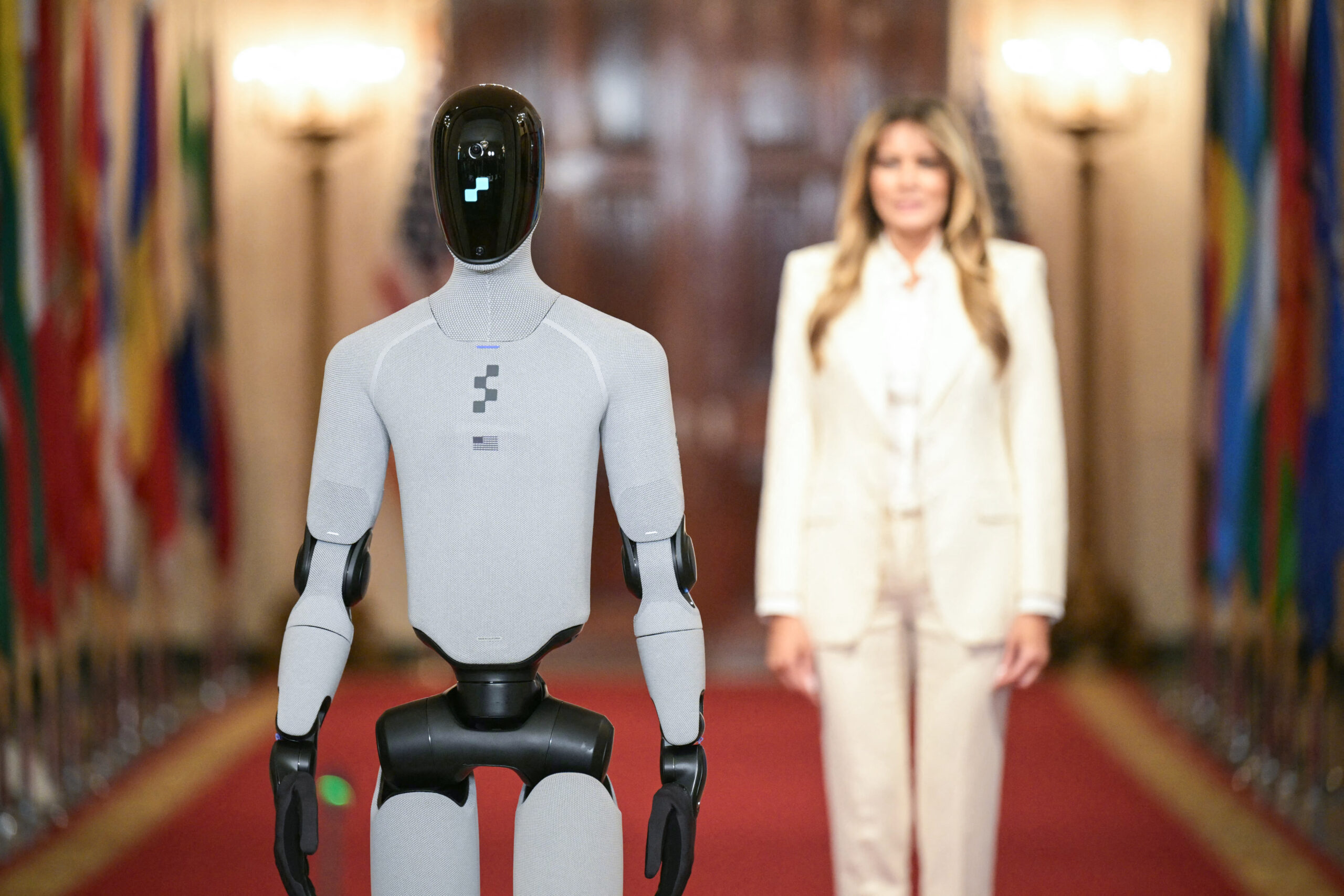 A humanoid robot stands in the hallways of the White House east wing. Melania Trump stands out of focus behind it.