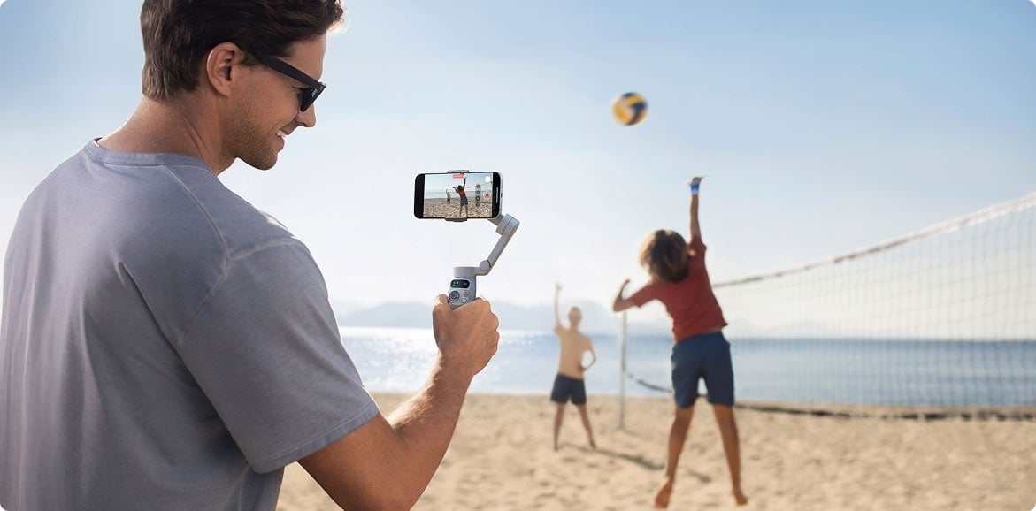 a person holds up the DJI Osmo Mobile 7 to film people playing volleyball on a sunny beach