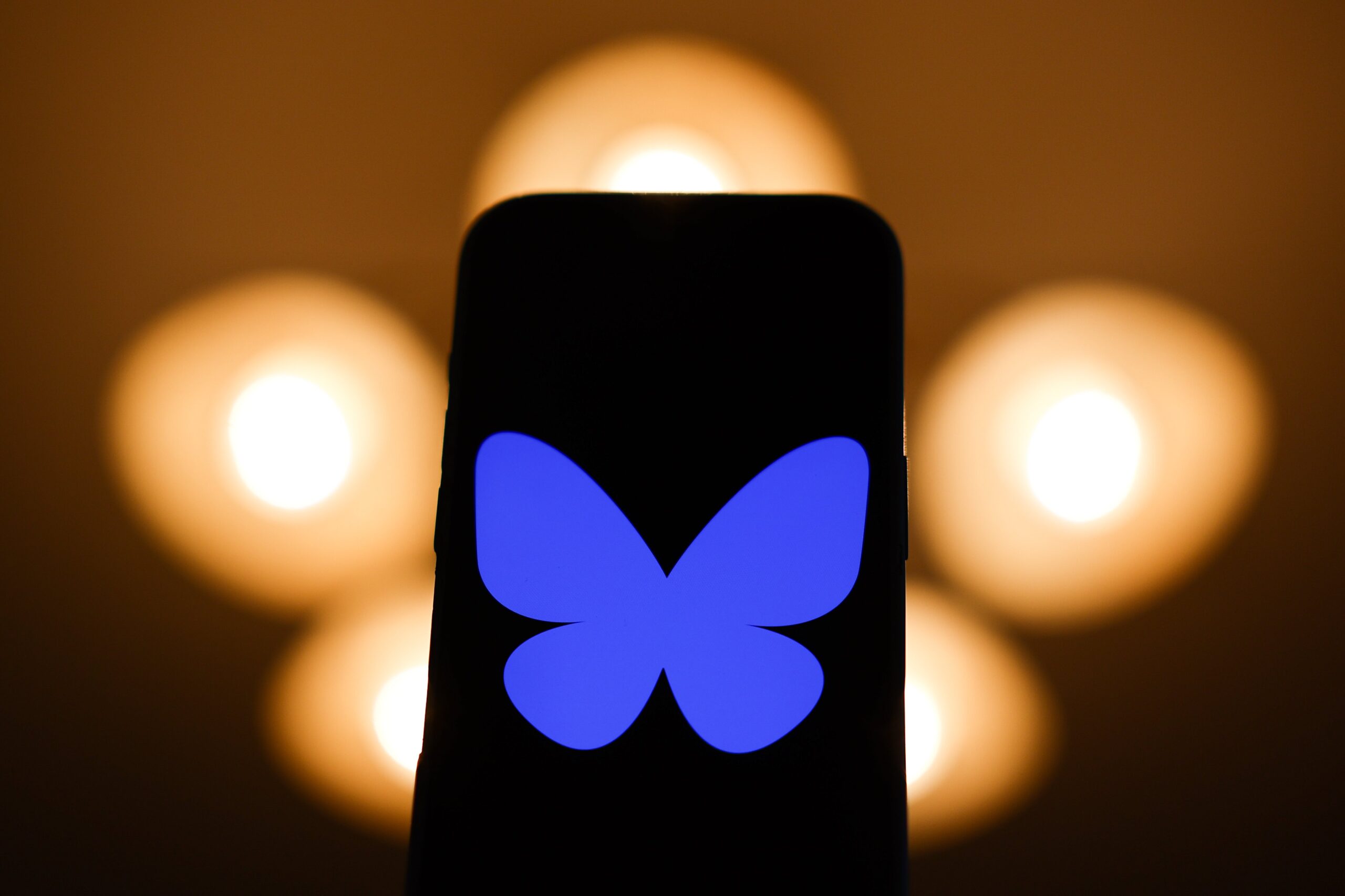 A phone in front of blurred shows a large Bluesky logo.