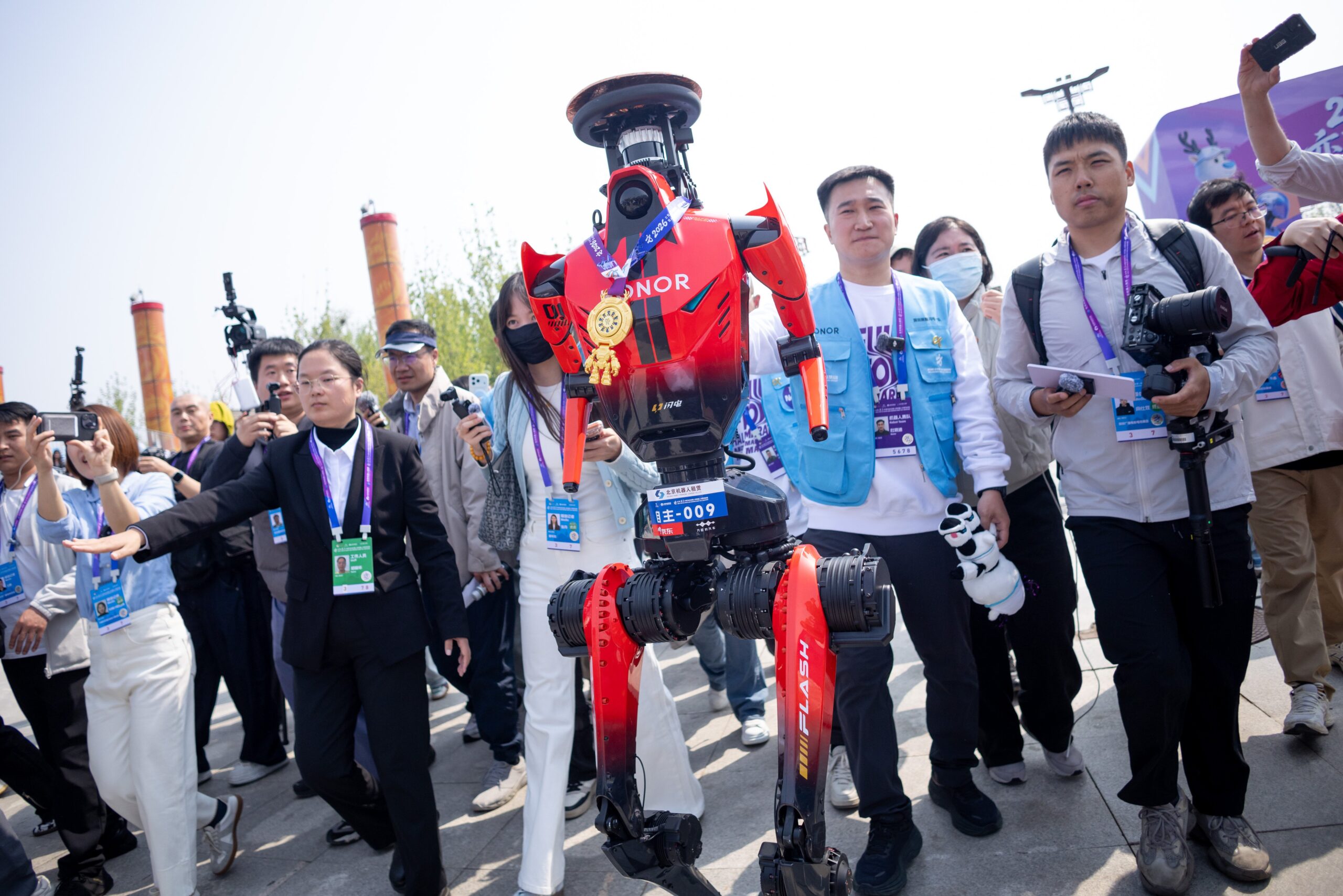A red, humanoid robot wearing a gold medal stands in front of a crowd of cheering humans. 